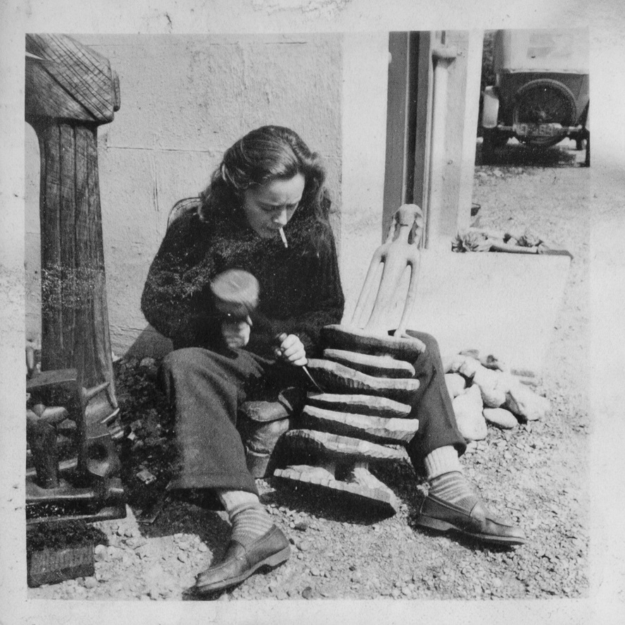Hilary Heron carving Lady of the Rocks circa 1953. Photo courtesy Hilary Heron Estate | Hilary Heron: A Retrospective | Friday 24 May  – Monday 28 October 2024 | IMMA | Image: Hilary Heron carving Lady of the Rocks circa 1953. Photo courtesy Hilary Heron Estate | black-and-white photo of the artist with mallet and chisel in hand, cigarette in mouth, working on a wooden idol-like sculpture which looks to be a bit over a metre high but is positioned between the artist’s legs, leaning against the left one; we’re outside; there’s the rear of an ancient jalopy in the background, a doorway almost seen from the side, the hint of a house, and a hint of 3D artworks too, to the artist’s right 