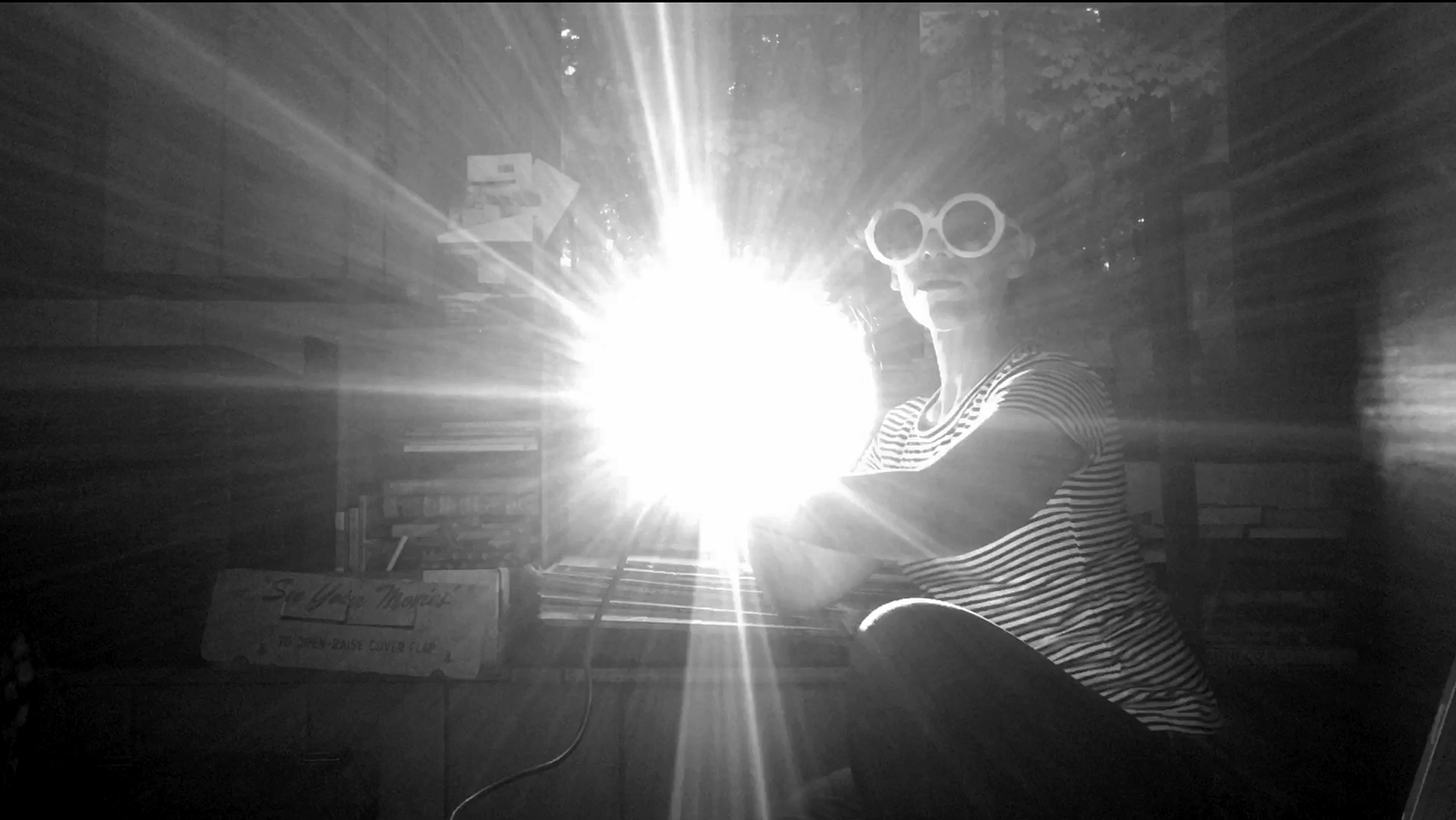 Gestures for a Plague Season or Goodby Earthworks, 2020, Nicole Antebi | Nicole Antebi: BORDER / RIVER / MOVER / BREATHER | Tuesday 7 May  – Thursday 9 May 2024 | NCAD Gallery | Image: Gestures for a Plague Season or Goodby Earthworks, 2020, Nicole Antebi | in a space that has plenty of books in the background, someone is possibly at a film projector; wearing large white, round-rimmed glasses, a striped top; in the centre of the black-and-white photo is a blaze of light, from the possible projector 