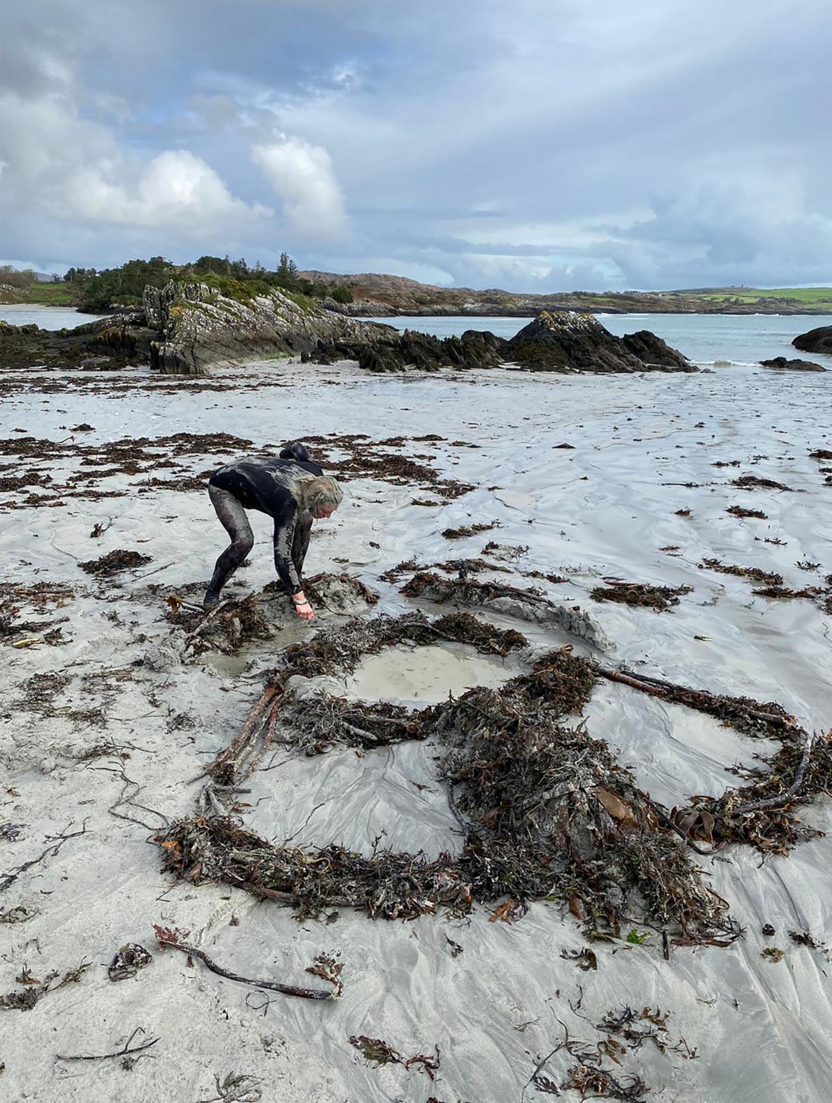 BAVA final year student Hammond Journeaux working on Sherkin Island | BA Visual Art Graduate Exhibition 2024 | Saturday 18 May  – Saturday 15 June 2024 |  | Image: BAVA final year student Hammond Journeaux working on Sherkin Island | photo of the artist arranging seaweed – in the outline of a bell or a skirt or… – on a beach; the artist, wearing what may be a black wetsuit, is leaning forward three or four metres from the camera, touching something towards the top of the bell / skirt; farther away we see typical craggy, barncle-encrusted rocky seaside outcrops, then sea, then trees and land, then a busy, cloudy sky