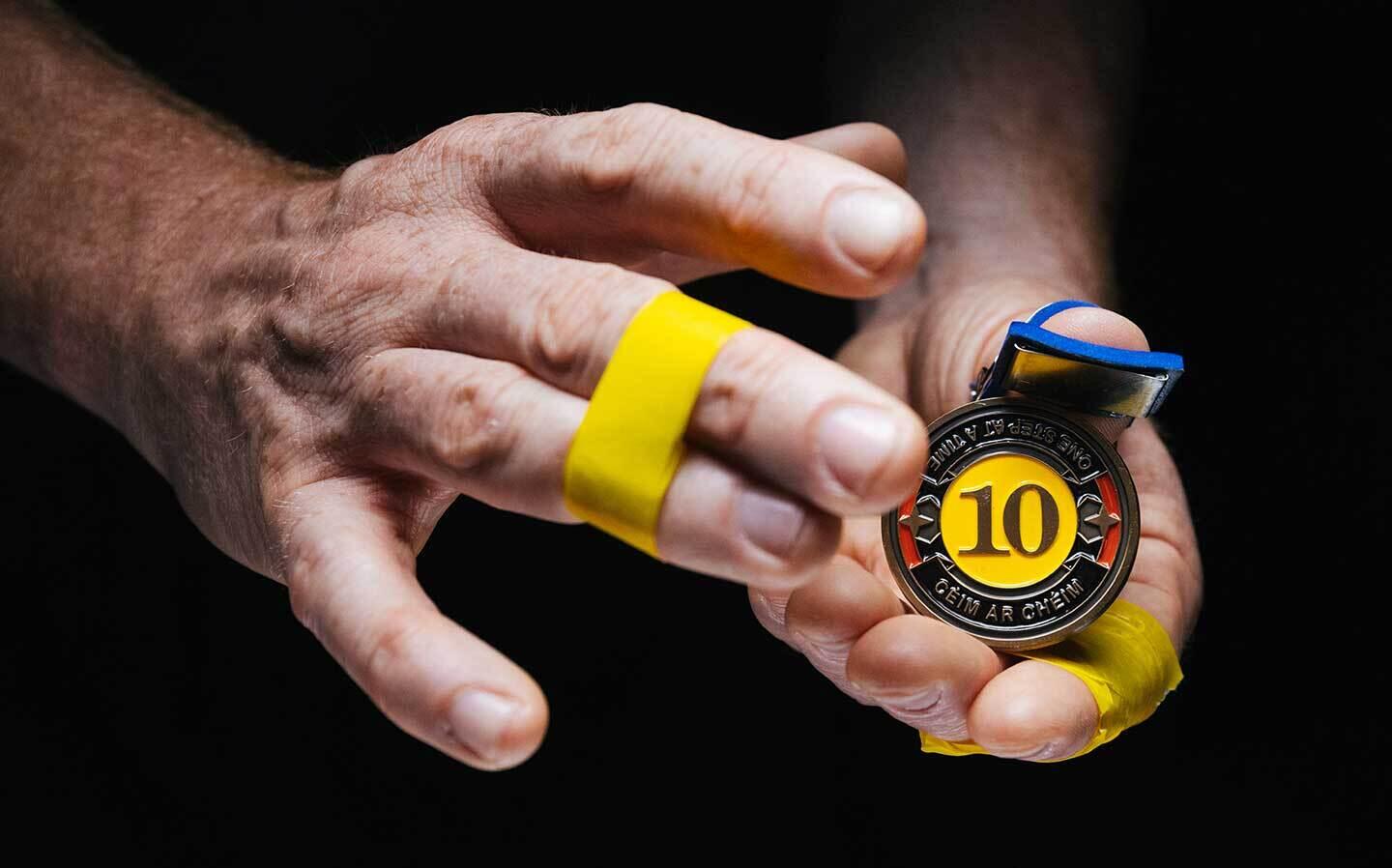 Brognon Rollin: Templates of Chance | Friday 7 June  – Saturday 31 August 2024 | Rua Red | Image: photo or video still of two hands, one holding a medal; the medal has the number 10 in the middle, on a strong yellow, circular background; the text in a band around this circle reads ‘ONE STEP AT A TIME | CÉIM AR CHÉIM’; the background is black; there is bright yellow tape binding fingers on either hand – the middle and ring finger on the right hand, the index and middle on the left hand 