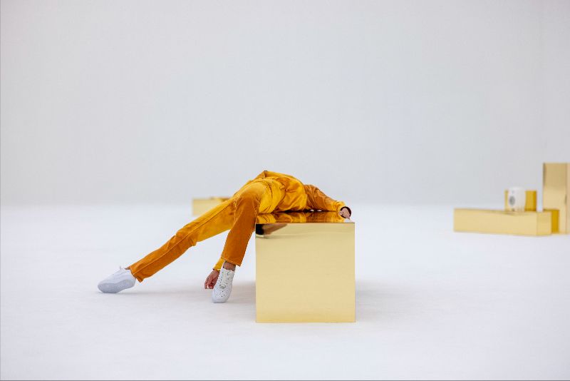 Maria Hassabi: White Out, 2023. Installation view, Tai Kwun Contemporary Hong Kong, Oct 12 – Nov 26, 2023. Performer: Marah Arcilla. Photo, Thomas Poravas. Courtesy the artist. | Take a Breath | Friday 14 June 2024  – Monday 17 March 2025 | IMMA | Image: Maria Hassabi: White Out, 2023. Installation view, Tai Kwun Contemporary Hong Kong, Oct 12 – Nov 26, 2023. Performer: Marah Arcilla. Photo, Thomas Poravas. Courtesy the artist | photo of a setting with various rectangular-shaped boxes, some at least apparently with a golden, reflective surface; otherwise the scene is in a white space; we see one of the boxes head-on, in the centre of the photo horizontally but around the bottom third of the image; a figure in an orange get-up is lying on the box, and we see only torso, arms and legs – though of the arms really only the hands; the figure is wearing white trainers 