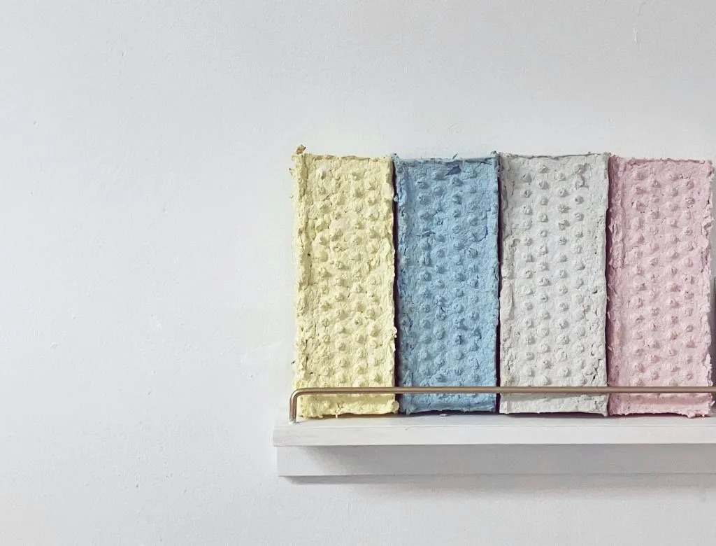 Ciara Rodgers, Folly | Façade,  | Saturday 8 June  – Saturday 13 July 2024 | GOMA Gallery of Modern Art | Image: photo of four brick-shaped objects on a shelf; each is effectively identical, except for the colour – pastel yellow, then blue, then greyish white, then pink; the ‘bricks’ are in vertical orientation, and there is a retaining metal (?) bar stretching across in front of them; it’s hard to make out what the ‘bricks’ are made of – possibly papier mâché 