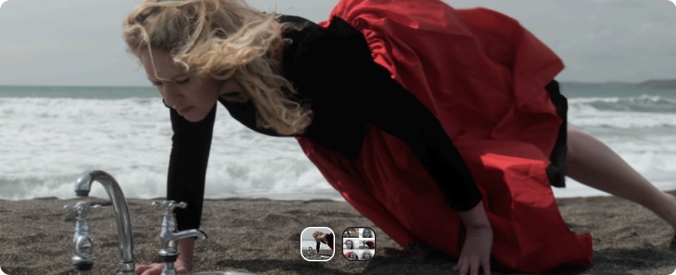 Etaoin Melville: Silent Voices – Cork Harbour Festival | Saturday 1 June  – Monday 10 June 2024 | Triskel Arts Centre | Image: film still; we are on a beach, with the sea frothing in the background and greyish sand closer to us; it’s clearly somewhat sunny; to the left in the image there is a stainless-steel kitchen sink embedded into the sand, with the two taps, faucet and draining board above ground; a figure with flowing long blond hair – caught by the wind – and wearing a black top and scarlet billowing skirt fills most of the central area of the image, almost looking like they are doing a one-handed push-up over the sink 