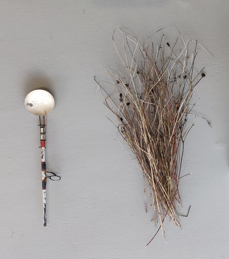 Stuart Cairns: Tideline Spoon and Spray; courtesy College Lane Gallery | Stuart Cairns: Hinterlands | Saturday 25 May  – Sunday 14 July 2024 | College Lane Gallery | Image: Stuart Cairns: Tideline Spoon and Spray; courtesy College Lane Gallery | photo of two objects on a wall; the one on the left is vertical and spoon-like, assembled with wire, what looks like the handle of a paintbrush which has been decorated in a somewhat Mondrian style + a circular curved blemished disk; the one on the right consists of maybe 100 or so dried stalks of long grass; some of the stalks have natural, black seedheads  