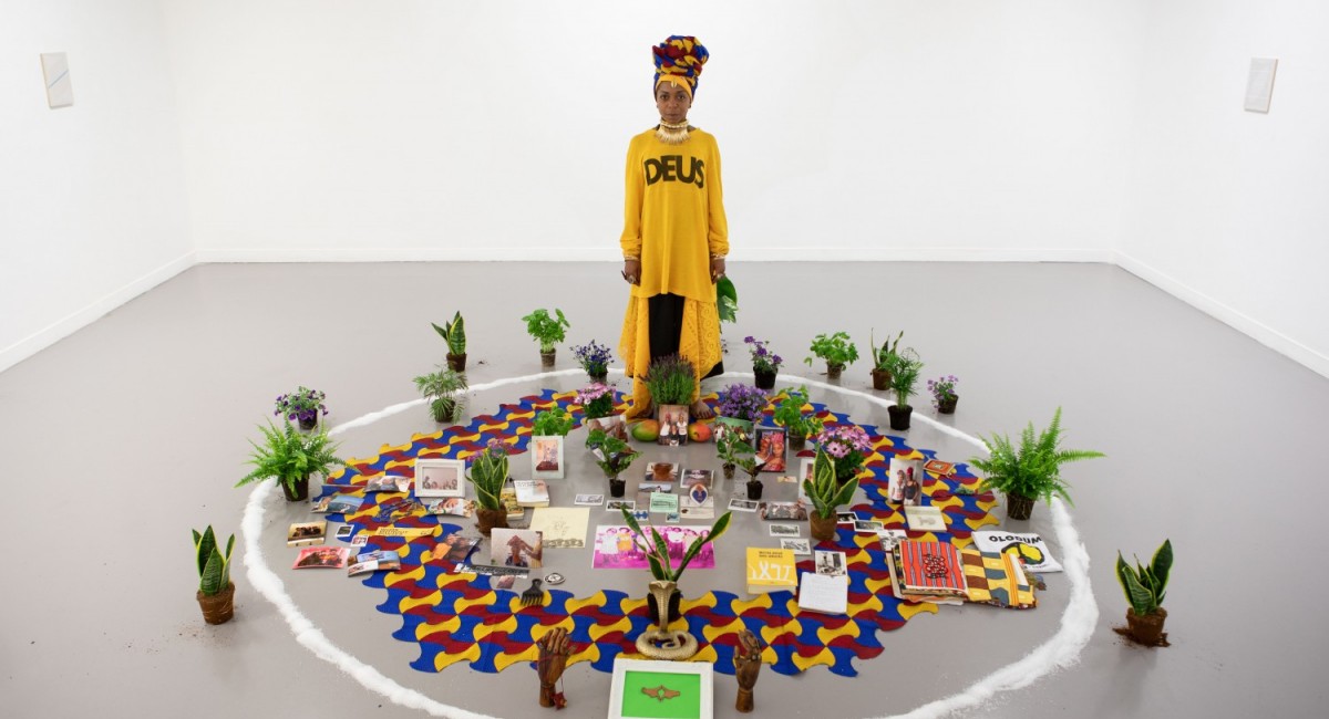 Thaís Muniz, Darling, Don’t Turn Your Back On Me (still), 2021. Courtesy of the artist | Thaís Muniz: Rites of Care, Curse & Comfort | Saturday 13 July  – Saturday 19 October 2024 | SIRIUS | Image: Thaís Muniz, Darling, Don’t Turn Your Back On Me (still), 2021. Courtesy of the artist | a figure, probably the artist, is standing in a gallery space – white walls with possibly a small, mostly white artwork haning on two of the walls – and grey floor; the figure is inside a white circle on the floor which is about three metres in diameter and apparently drawing using plentiful amounts of salt or sugar; the figure, dressed in a yellow / mustard long-sleeved outfit that reaches from shoulder to floor and wearing a complicated headdress that may be a basket of some sort, with possibly fruit in it; inside and around the circle we see many potted plants, of the fern or aloe vera variety, for the most part; inside the circle there are other objects, including two wooden hand poking from the floor, a hexagonal pattern in red, blue and yellow curved tiles, books, framed photos, possibly mangoes, other things 