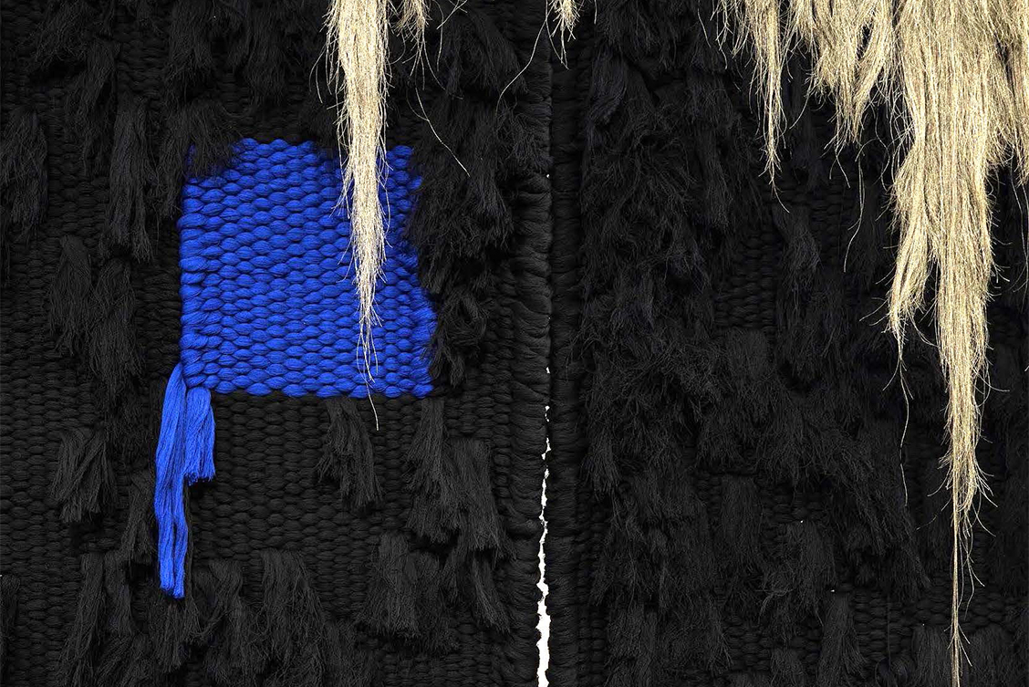 Emily Waszak: We Speak Through Worlds (detail), 2023 | Thresholds to the Unseen | Saturday 7 September  – Friday 1 November 2024 | Solstice Arts Centre | Image: Emily Waszak: We Speak Through Worlds (detail), 2023 | photo of a detail of a mixed-fabric work; most of the fabric is woven black, though there is a woven square that is a strong blue; ends (probably) of the woven black fibre hang from the black material at random points, as does the blue material dangle down from the left edge of the blue square; we see to see a slight vertical split between two parts of the black material; and hanging down from the top of the photo, we see long, shaggy bán hairs of what may be wool 