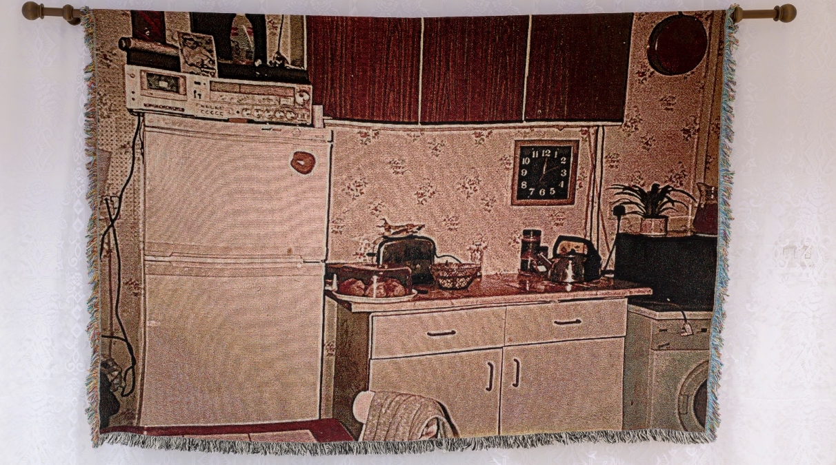 Michelle Malone, The Kitchen, 26 Cloonlara Crescent, 2022, digitally woven tapestry depicting Malone family council house kitchen in the late 80s, 244 x 122 cm, Image courtesy of the artist. | Wilton Park Studios 2019 to 2023 | Friday 6 September  – Sunday 20 October 2024 | Royal Hibernian Academy | Image: Michelle Malone, The Kitchen, 26 Cloonlara Crescent, 2022, Digitally woven tapestry depicting Malone family council house kitchen in the late 80s, 244 x 122 cm, Image courtesy of the artist. | this is a photo in woven form; there is a pinkish cast over the result, presumably from the age of the photo; the tapestry is depicted hanging on a wooden crossbar of the type that holds domestic curtains; in the photo; there is not much that is difrent from today – a fridge, toaster, electric kettle, formica top, edge of a washing machine, even what looks like a microwave; the one thing you’d be very unlikely to find nowadays is the fancy casette-player + tuner, which is sitting on top of the fridge 
