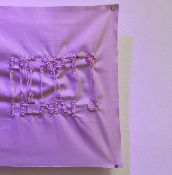 Chloe Austin: Living but a Day: Texere | Saturday 14 September  – Sunday 13 October 2024 | GOMA Gallery of Modern Art | Image: the illumination for this photo appears to be purple, so the textile we see is possibly white in daylight; it appears to hang on a wall, tacked to a support such as a wooden subframe, perhaps; it is casting a pale yellowish shadow; the textile, which is cropped out of the photo on the left, looks like cotton or linen with white (?) threads sewn into it, making tall shapes that are somewhat letter-like 