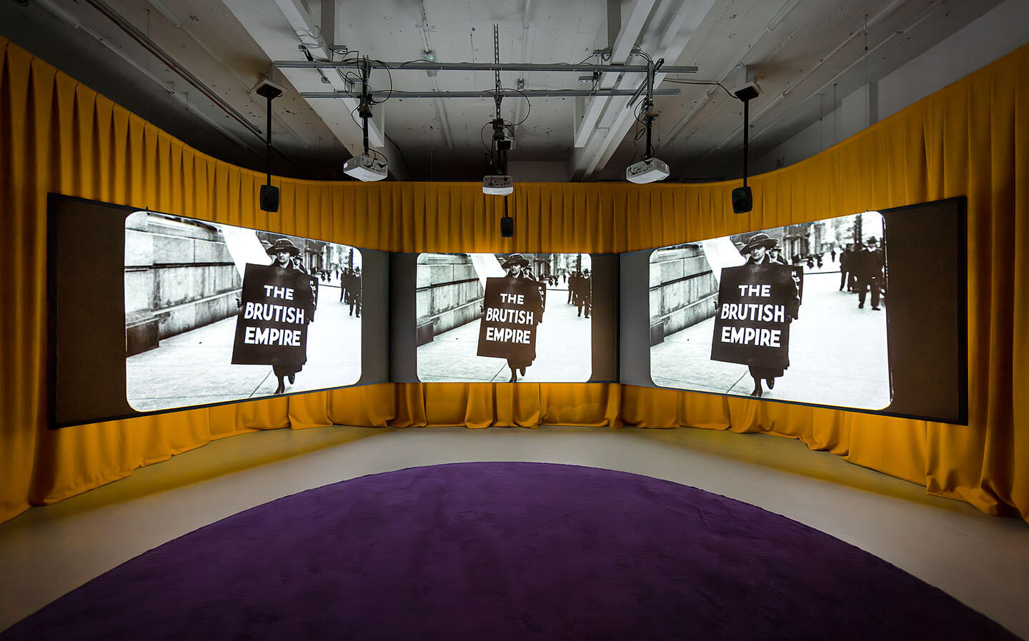 Marianne Keating, An Ciúnas / The Silence, 2023. Installation view, The Showroom. Photograph: Dan Weill | Marianne Keating: An Ciúnas / The Silence | Friday 11 April  – Sunday 8 June 2025 | Limerick City Gallery | Image: Marianne Keating, An Ciúnas / The Silence, 2023. Installation view, The Showroom. Photograph: Dan Weill | in this photo we see three screens hanging in front of a mustard-orange curtain; the floor is grey, but it has a purple, curving carpet on it; the image on each screen is the same: a black-and-white photo of a protester wearing a hat, overcoat, boots (?) and a large sandwich-board which is black with three words in white non-serif capitals on it: \