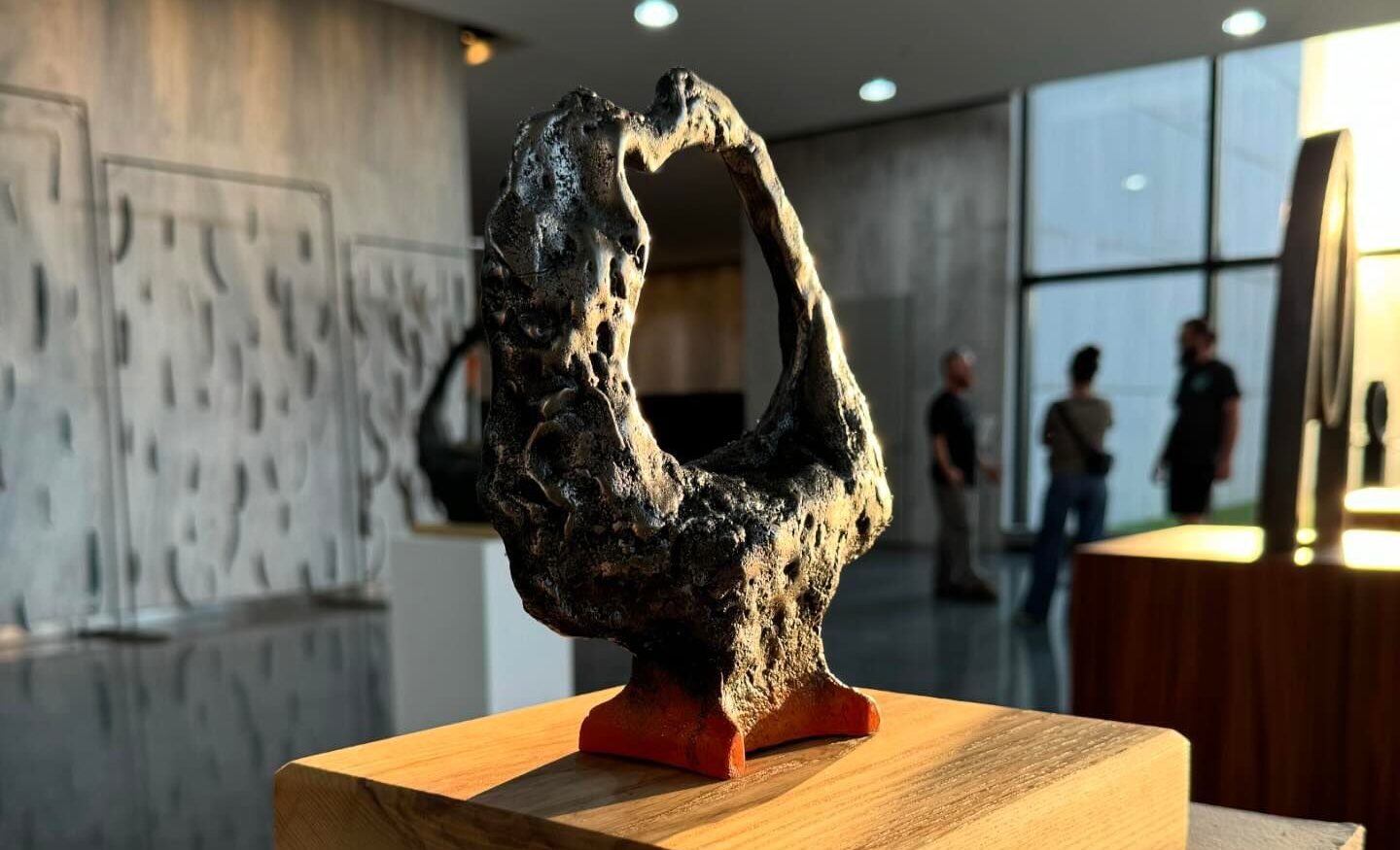 James L Hayes, The Tucson Anvil, 2024, Cast iron, American white ash & spray primer | Material Laboratories: Metal Fest | Tuesday 1 October  – Saturday 5 October 2024 | National Sculpture Factory | Image: James L Hayes, The Tucson Anvil, 2024, Cast iron, American white ash & spray primer | photo of the artwork in a large space with other sculptural pieces (maybe the Glucksman in Cork?); there are blurred figures to the right of the photo, and a few out-of-focus artworks; the 'anvil' has a base (which is orange) that is presumably like that of an anvil, but sprouting from it, so to speak, is something that looks like a pocked metal meteorite in the rough shape of a vertical circle; there is in fact a Tucson meteorite that looks very like this 