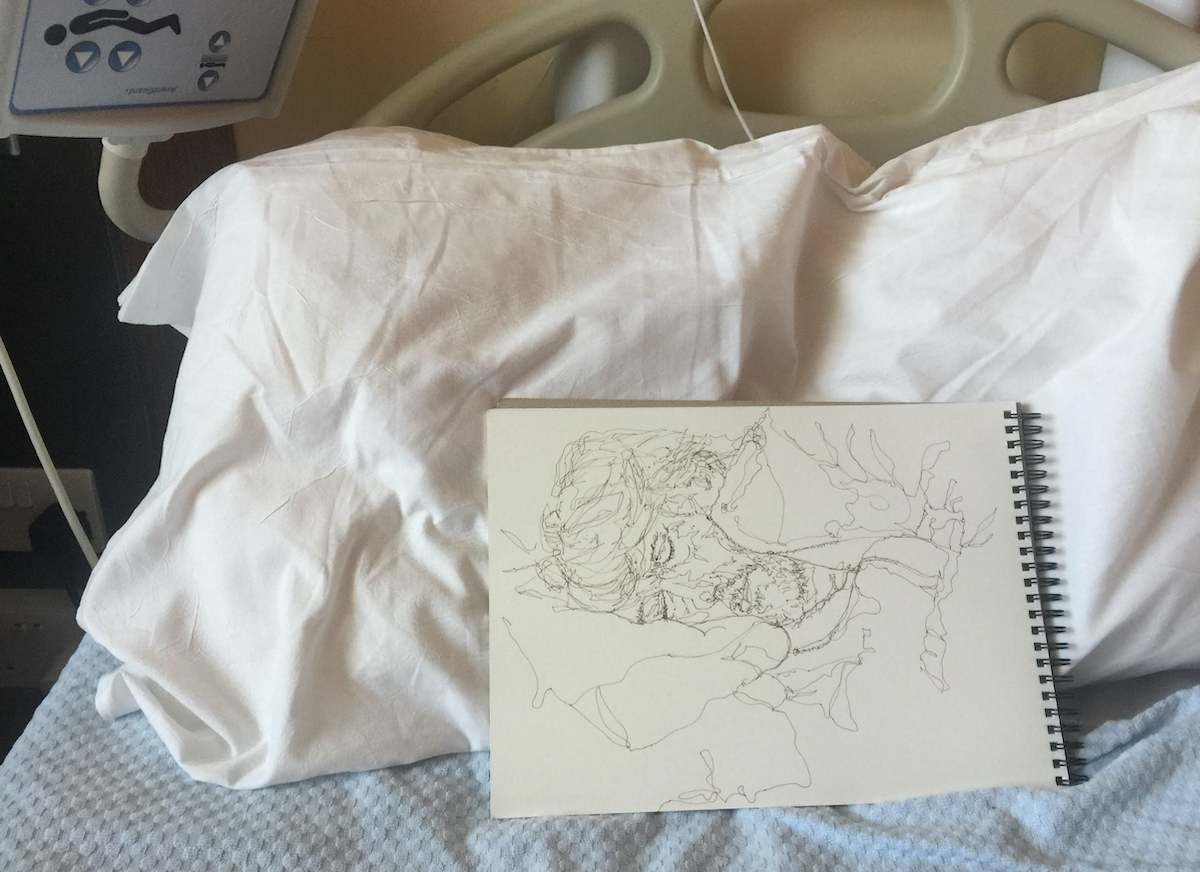 John Kelly & Christina Todesco-Kelly: A Time Otherwhere | from Friday 20 September  |  | Image: photo taken in a hospital setting – we see a pillow against the head of the bed, and top-right the device that nursing staff will press to raise or lower parts of the bed; it is upside-down; the pillow is white, and against it rests a sketchbook open to a page on which Christina Todesco-Kelly has drawn John Kelly as he was sleeping; it’s a head-and-shoulder study purely in line; he is on his right side, facing the artist, and we see his long unruly hair and bearded face 