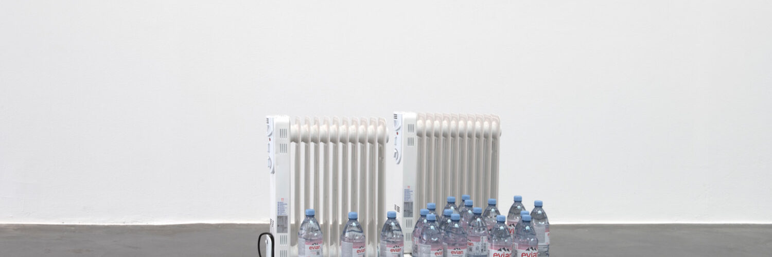 Installation shot of Trap Piece 7 BPA Sneckdown, Espacio de Arte Contemporaneo de Castellon 2023 EACC Photo Roberto Ruiz Arguedas Plastic water bottles and heater | Lucie McLaughlin, Francis Whorrall-Campbell, Ebun Sodipo: REHEARSING BODIES | Saturday 5 October  – Tuesday 5 November 2024 | CCA | Image: Installation shot of Trap Piece 7 BPA Sneckdown, Espacio de Arte Contemporaneo de Castellon 2023 EACC Photo Roberto Ruiz Arguedas Plastic water bottles and heater | photo showing two old-wall-radiator-style electric radiators standing behind 18 unopened bottles of water which are not arranged in any particular fashion; they have pale-blue caps; the floor (of which we see very little) is grey, and the wall behind is white 