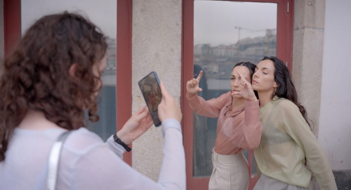 Lisa Freeman, Approx 1 Second of a Sweet Kiss, still, 2024 | Lisa Freeman: Approx. 1 Second of a Sweet Kiss | Saturday 14 September  – Saturday 12 October 2024 | SIRIUS | Image: Lisa Freeman, Approx 1 Second of a Sweet Kiss, still, 2024 | still image, showing three figures; the one on the left is evidently taking a photo with a mobile phohne, to the right and slightly farther away the other two are posing up against each other while facing the camera; the one on the left is making a sort of framing sign with her fingers; behind there are two lare reflective windows, and we see some sort of town- or cityscape, presumably the 'unfamiliar city in the south of Europe' from across a small stretch of water; all three figuyres have dark hair, probably long in each case; it looks as though all three may be wearing the same type of blouse, though in different pale colours; it must be a warm-enough day \