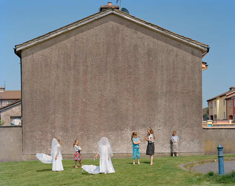 Doug DuBois, My Last Day at Seventeen: Aisling and Jemma’s First Communion, 2011, IMMA Collection: David Kronn Collection, 2024, © Doug DuBois | David Kronn Photography Collection | Monday 28 October 2024  – Sunday 26 January 2025 | IMMA | Image: Doug DuBois, My Last Day at Seventeen: Aisling and Jemma’s First Communion, 2011, IMMA Collection: David Kronn Collection, 2024, © Doug DuBois  | photo taken in a housing estate, probably; facing us is the full gable end of a house which seems to be adjacent to a green space; close to the gable is a group of kids; two of them are in first-communion clothes – bridal wear, effectively, including veils over their faces (instead of tossed back oder their heads); both also have white umbrellas, which they are holding open but downwards at their sides; the other kids are in everyday clothes; it’s sunny with a clear blue sky