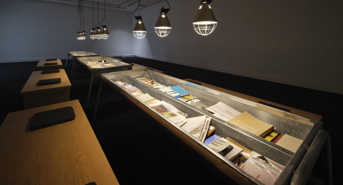 Deirdre O'Mahony, Feeder, Royal Hibernian Academy, 2022. Photo: Ros Kavanagh | Deirdre O’Mahony: Feeder | Saturday 12 October  – Saturday 2 November 2024 | SIRIUS | Image: Deirdre O'Mahony, Feeder, Royal Hibernian Academy, 2022. Photo: Ros Kavanagh | installation shot of what is probably reading material associated with the exhibition; the material is in what looks like cattle troughs, three of them lit from above by what may also be farm-related lights; along both sides of the troughs are wooden benches, each with two cushions, presumably for potential readers to sit 