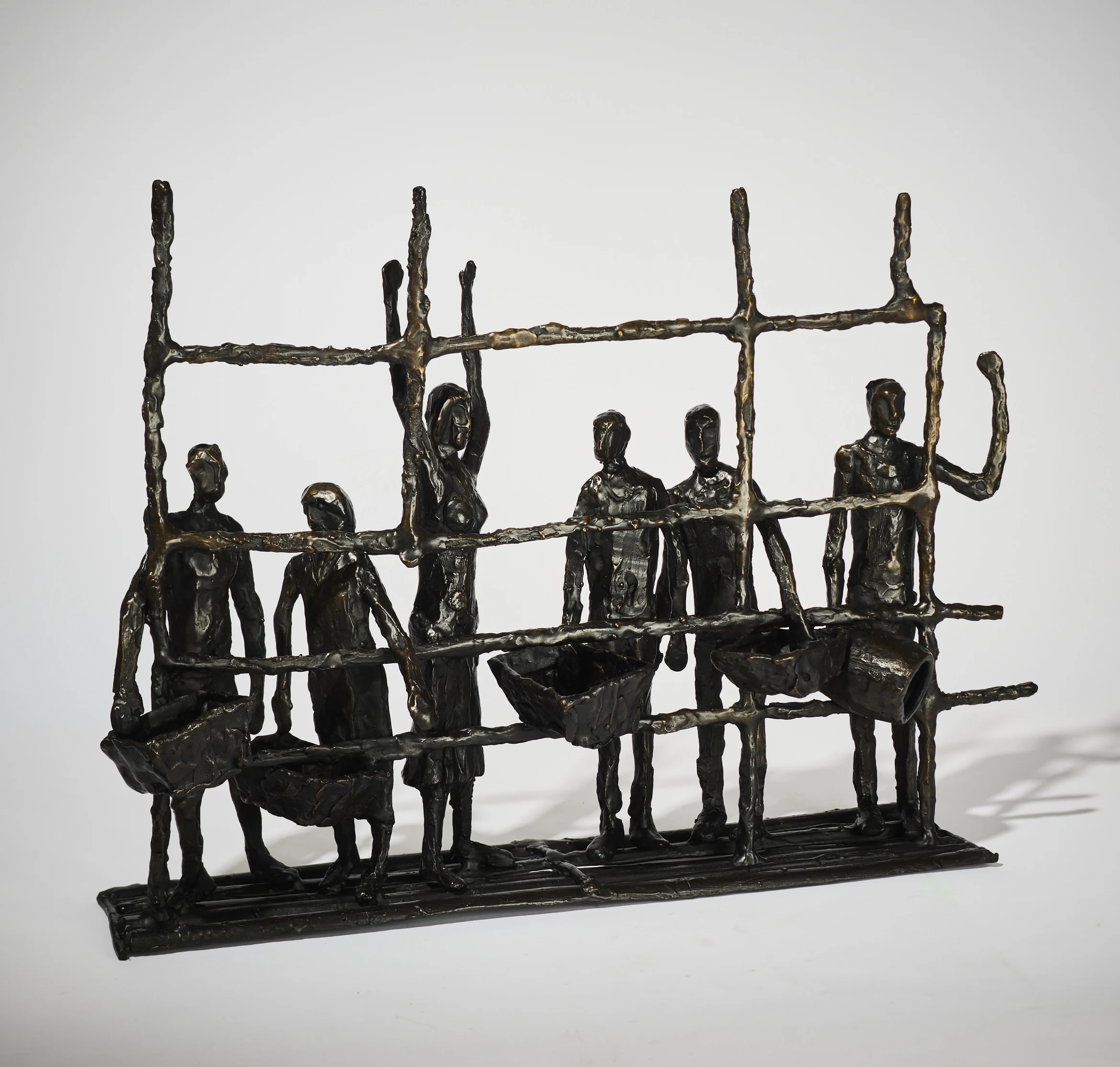 John Behan: Walking into Eternity | Thursday 24 October  – Saturday 16 November 2024 | Solomon Fine Art | Image: photo of a bronze artwork in white space; we see six figures, five of them carrying large, boxy baskets; the one without a basket is raising both arms; they are stuck, apparently, at the other side of a barrier, indicated by a horizontal and vertical bars of bronze, for horizontal, four vertical 
