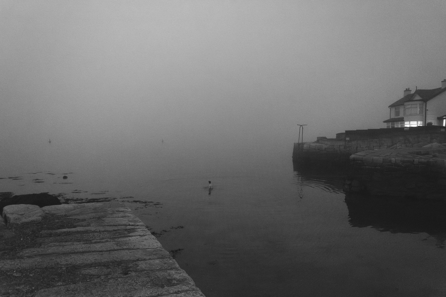 Róisín White: Safe Harbour | Friday 8 November 2024  – Sunday 19 January 2025 | Municipal Gallery | Image: greyscale photo or video still, taken, at a guess, in the Sandycove part of Dún Laoghaire / Rathdown; we see hints of pier left and right, and that peculiar suburban house that somehow got built on the pier towards the right edge of the image; in the house there is a light on in what may be the living-room, making the bay window glow; apart from light from the house, things are gloomy – it’s a very dull day, or dawn or dusk; there is a thick fog, but we can see a swimmer – who is probably standing – in the very calm seawater, in the centre of the image; there may be two other swimmers farther out, or those may be buoys 