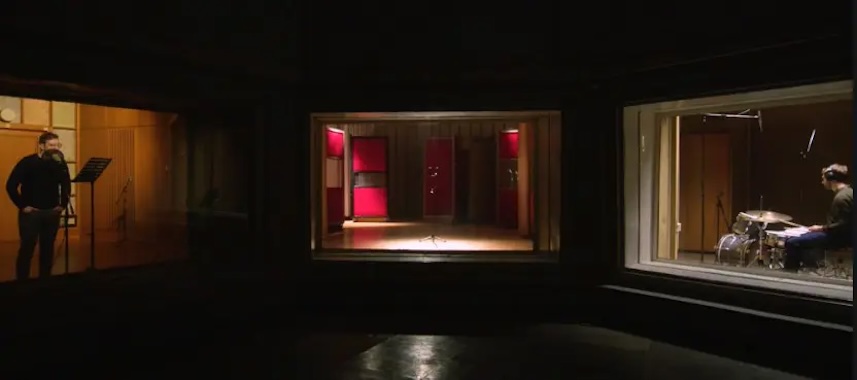 Lawrence Abu Hamdan, Walled Unwalled, HD video, stereo sound, 2018. Collection & image © Hugh Lane Gallery. | Lawrence Abu Hamdan: Walled Unwalled | Thursday 3 October 2024  – Sunday 18 May 2025 | Hugh Lane Gallery | Image: Lawrence Abu Hamdan, Walled Unwalled, HD video, stereo sound, 2018. Collection & image © Hugh Lane Gallery | the video shows what seems to be a view into three recording studios at the same time, through a very large glass window, seen from a dark space; the studio on the left has one figure standing, talking or singing in front of a microphone; the room through the window straight ahead is empty; there is some of stand in the middle of the room, possible a microphone stand, and we see red doors or soundproofing; through the window on the right we see a drummer at a drum kit 