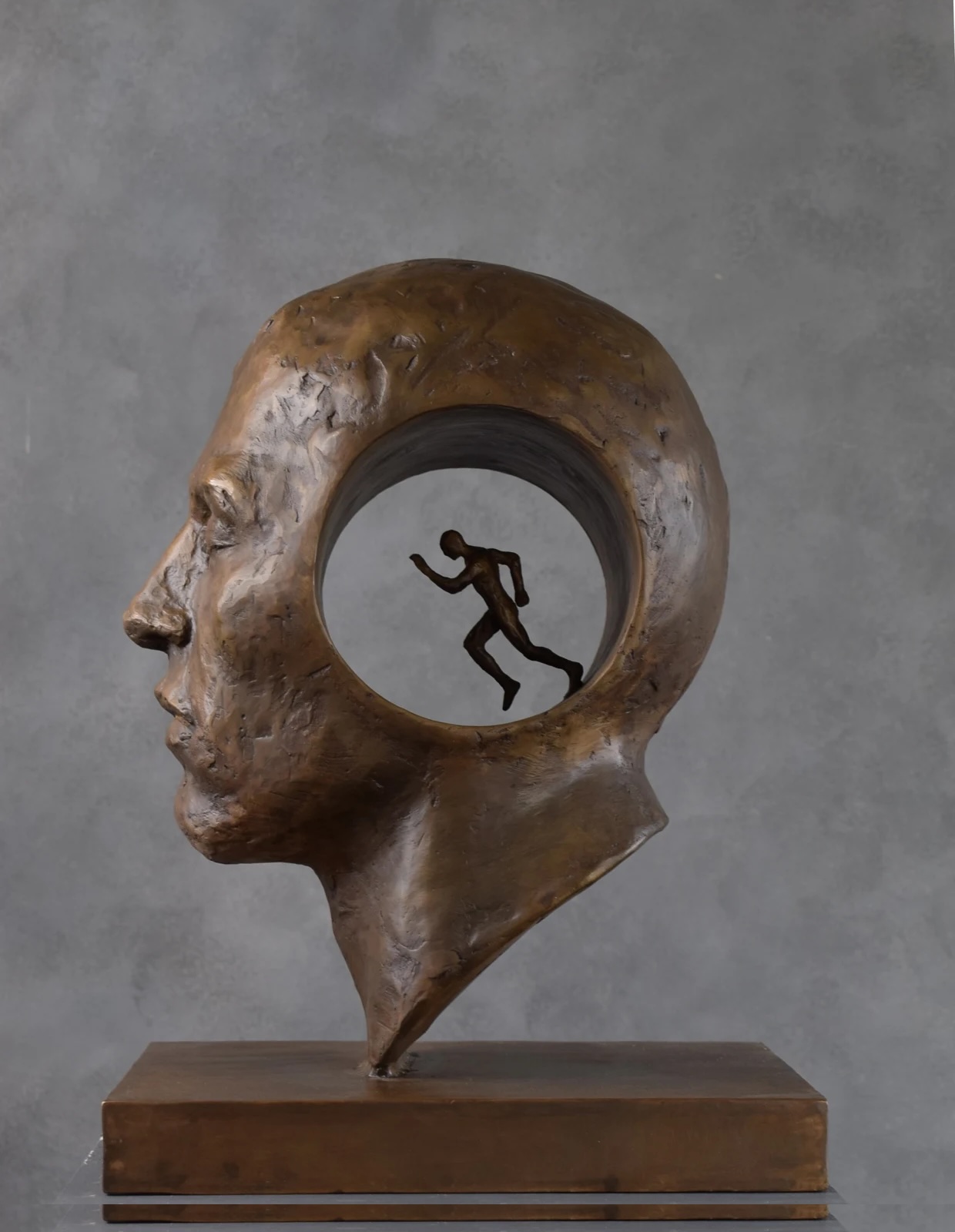 Mark Rode: Perpetual Motion, bronze, height 45 cm | Mark Rode: Inner Selves | Thursday 7 November  – Sunday 24 November 2024 | Gormleys Fine Art, Dublin | Image: Mark Rode: Perpetual Motion, bronze, height 45 cm | in the photo of the work, we see a head in bronze looking towards our left, in profile and realistically portrayed; going right through the head is a large round hole and in it we see a figure running, heading to our left; the background in the photo is a cloudy, uniform grey; daylight is reflected off the bronze 