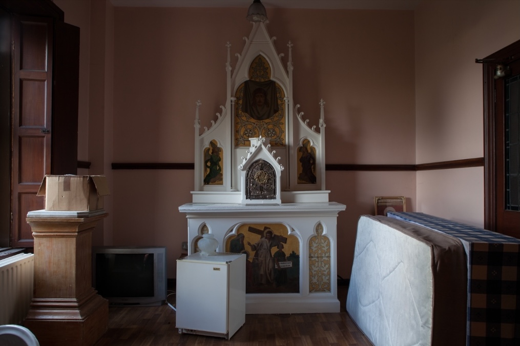 Catriona Leahy, Rupture II, 2024, Graphite drawing and transfer print on Shiohara Japanese Paper, 60cm x 26cm, Image courtesy of the artist | Pauline Rowan: Under a Vaulted Sky | Saturday 23 November 2024  – Sunday 2 February 2025 | Royal Hibernian Academy | Image: Pauline Rowan, Tabernacle under a Vaulted Sky, 2024, Image courtesy of the artist. | photo taken in what is presumably a side room in a convent; we see a small but ornate altar, Gothic style, with various religious themes in gold-leafed paintings among the marble; but we also see that it is a storeroom – a small fridge with a vase on it in front of the altar, two mattresses apparently blocking a door to the right, a cardboard box on a dusty, chunky wooden plinth in the tall window to the left, of which we only see the dark wooden shutters on its right side; the walls of the room are painting pale salmon pink; it’s gloomy 