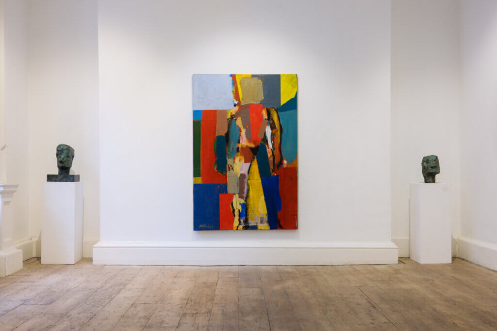 Michael Kane, Brian Bourke. Installation view, Taylor Galleries. Photo by Conor Horgan. | Group Show: 24 @ TG | Friday 8 November  – Thursday 12 December 2024 | Taylor Galleries | Image: Michael Kane, Brian Bourke. Installation view, Taylor Galleries. Photo by Conor Horgan. | photo of the interior of the gallery; immediate facing us, hanging on a white wall, is a large painting by Michael Kane; portrait orientation, strong bright colours and darker, partial outlines, suggesting a figure; to the right and left of the painting, on plinths tucked into matching alcoves, are two sculpted heads by John Behan, in a  greenish patina suggesting that they are bronze 