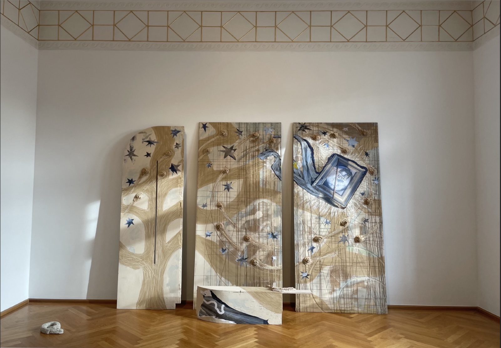 Alice Rekab, You Could Not Choose the Star You Were Born Under, 2022. Installation view, Museum Villa Stuck, Munich. Photograph: Jann Averwerser. Courtesy of the artist | Alice Rekab: Clann Miotlantach / Mythlantics | Saturday 23 November 2024  – Saturday 5 April 2025 | SIRIUS | Image: Alice Rekab, You Could Not Choose the Star You Were Born Under, 2022. Installation view, Museum Villa Stuck, Munich. Photograph: Jann Averwerser. Courtesy of the artist | in the photo we see three artworks leaning against a wall in front of us; there may also be two more artworks resting on the parqueted floor; light is falling on the works through a window, unseen, to the right; the walls of the room are very tall, and at cornice level there is a repeating geometrical pattern, possibly in gold paint, in a simple art-deco or art-nouveau style; each of the three artworks leaning against the wall appear to be painted on board, or possibly canvas; each is possibly two metres high, though two of them are narrow, about half as wide as they are high, and the third is even narrower; it is possible that the three panels are continuations of each other, as a sort of tree-like motif seems to join up between the panels, especially the two on the right; it is hard to make out what exactly is being portrayed in the muted browns on pale-tan surface, for the most part