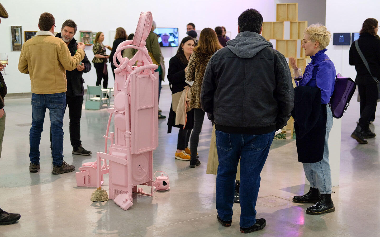 Borders: Rua Red’s Annual Exhibition | Friday 22 November 2024  – Saturday 25 January 2025 | Rua Red | Image: ‘Displacement and Belonging’ – Home, Rua Red Annual Open, 2023. Photo by David Reilly | photo of what is probably the opening of the annual exhibition a year ago; we see many people standing around chatting in a large gallery space; quite close to the camera is a human-size sculpture, which does not particularly resemble a human; it is painted a uniform pink and possibly constructed out of various domestic objects; one ‘leg’ of the sculpture ends in what looks more or less like a small mound of porridge; near another leg is a pink teapot with a hole knocked in it 