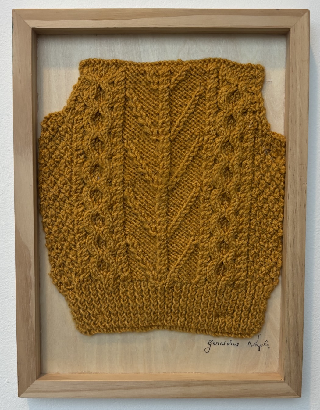 Geraldine Nagle: Seasons Through Stitch | Winter Light | Wednesday 6 November 2024  – Saturday 25 January 2025 | Uillinn: West Cork Arts Centre | Image: Geraldine Nagle: Seasons Through Stitch | photo of a knitted object in a wooden frame; cable-stitch-type knitting, but it’s not clear what the object is – maybe the back of a sleeveless sweater?; the colour is a earthy orange / yellow – raw umber? 