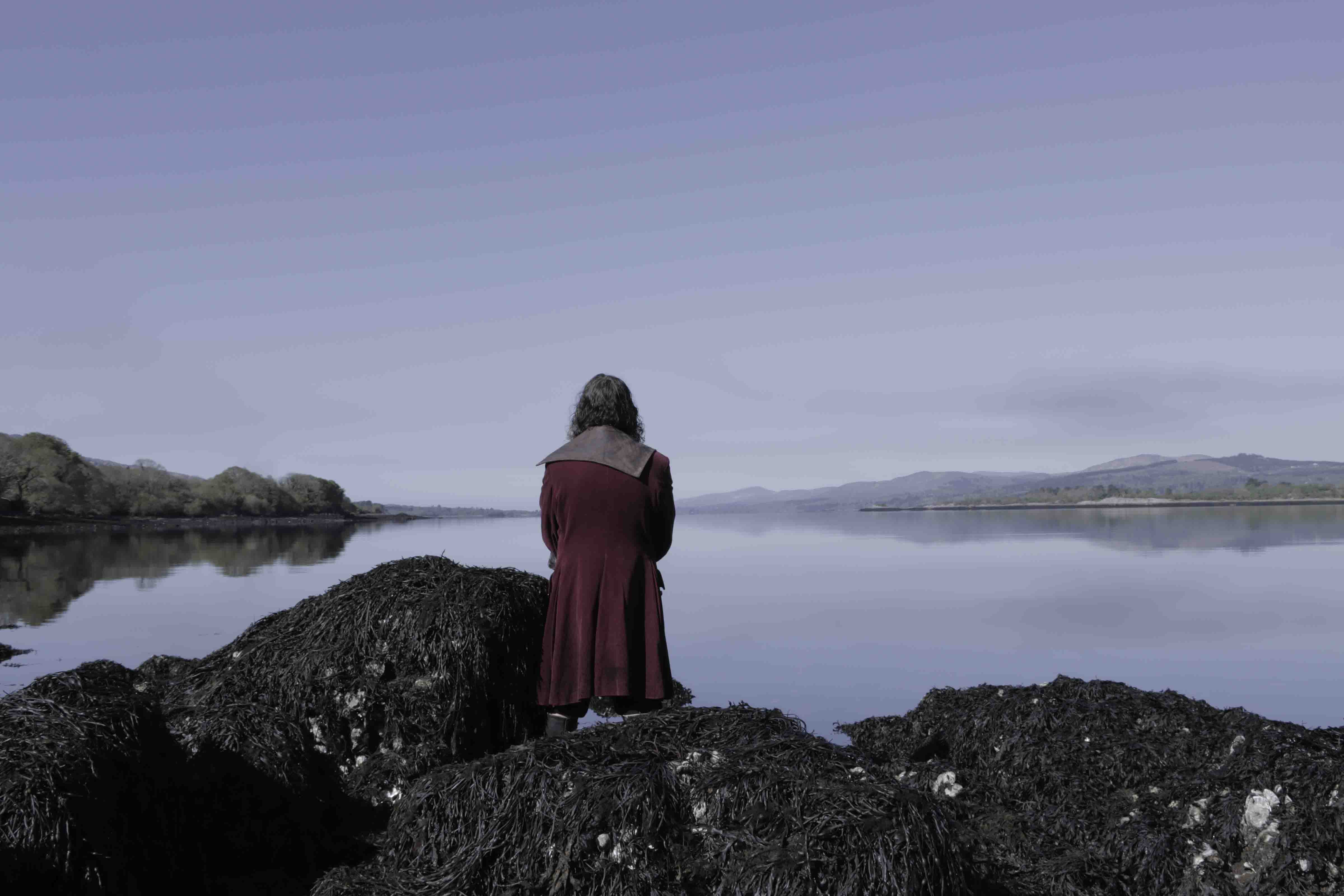 Myfanwy Frost Jones | water has no enemy | Thursday 12 December 2024  – Saturday 4 January 2025 | The LAB | Image: Myfanwy Frost Jones | in a photo or video still we see a figure looking out to sea, back turned to us; long dark hair, maroon coat (with hood?); the shoreline is rocky – rounded boulders covered in seaweed; it looks like a still, cold, almost cloudless day in winter; there are low hills in the distance across the calm water 