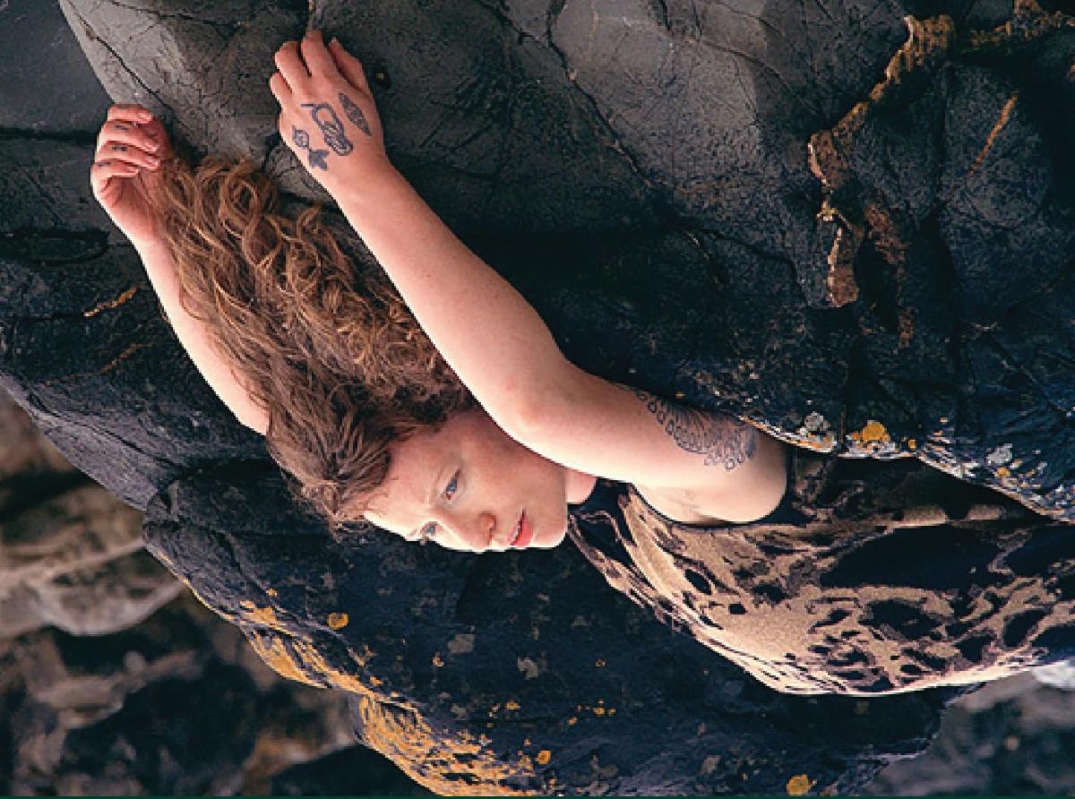 Caoimhín Gaffney: All At Once Collapsing Together | Saturday 11 January  – Saturday 15 February 2025 | Uillinn: West Cork Arts Centre | Image: photo of a pale-skinned figure with long brown, semi-curly hair lying across some dark, lichen-specked rocks, probably at the seaside, arms stretched back over the head and onto the rocks, just head, upper body and arms; the left hand, which is closest to us, bears three enigmatic tattoos, and there is a partial view of another on ntheleft shoulder; oh, and the whole image is upside-down  