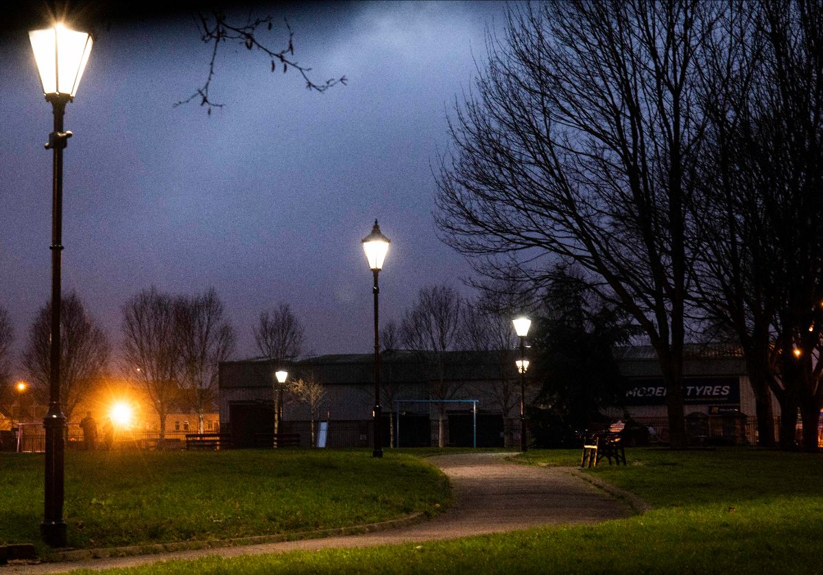 Maddie Leach: Evening Echo | Wednesday 1 January 2025 |  | Image: photograph, presumably of Shalom Park; very atmospheric, presumably taken at sunset; we see five old-fashioned black lampposts, each with a lit, white light on them, at the side of a gravelly path through the park; there is a bench on the right of the path, maybe five metres away; two figures seems to be standing in the background on the left, and behind them is a very powerful source of yellowish light – possibly the setting sun reflected in the window of a house – we see what seem to be a few houses at the edge of the park; there are trees i nthe park, bare of leaves 
