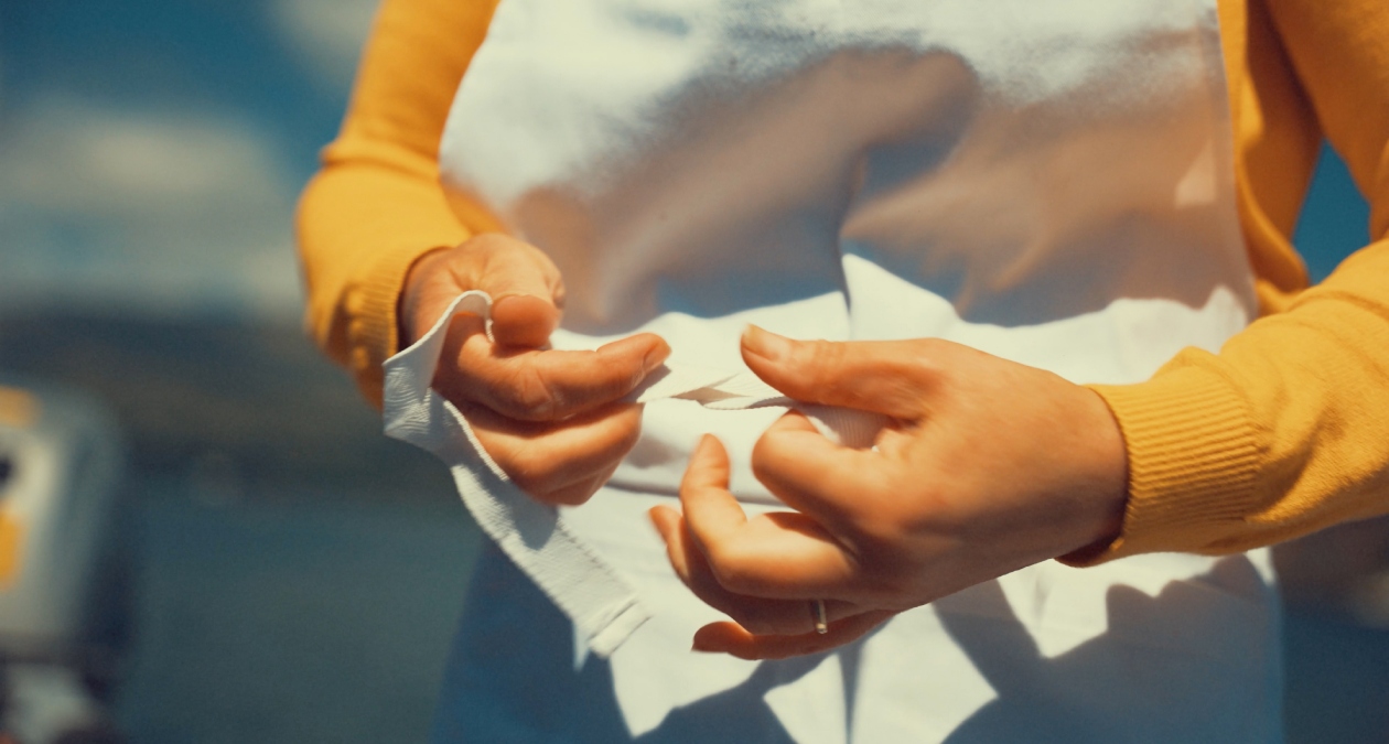 Mary Sullivan, The Fine Line, 2022, Video still, Photography Mickael Do Coutoa, Image courtesy of the artist. | Mary Sullivan: From the Inside Out and the Outside In | Friday 21 February –    Sunday 23 March 2025 | Royal Hibernian Academy | Image: Mary Sullivan, The Fine Line, 2022, Video still, Photography Mickael Do Coutoa, Image courtesy of the artist. | in the video still we see only torso to waist of someone in a bright orange top and white apron holding, it seems, a piece of white cloth which they are folding, judging by the stilled movement of the hands; behind, there is a blurred suggewstion of landscape – flat land and clouds; it is sunny 
