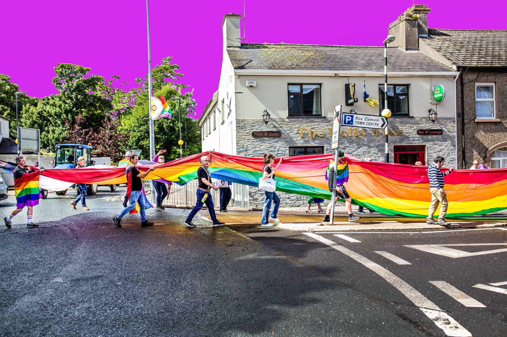 Jessica Weber Patterson: True Colours Shining Through | Thursday 13 February  – Thursday 27 March 2025 | Roscommon Arts Centre | the photo is a side view of march participants carrying a veeery long pride flag through the streets of Roscommon on a sunny day; we see trees and building in the background, and an LGBT+ flag is hanginging from a lamppost; the sky is a startling uniform puce 
