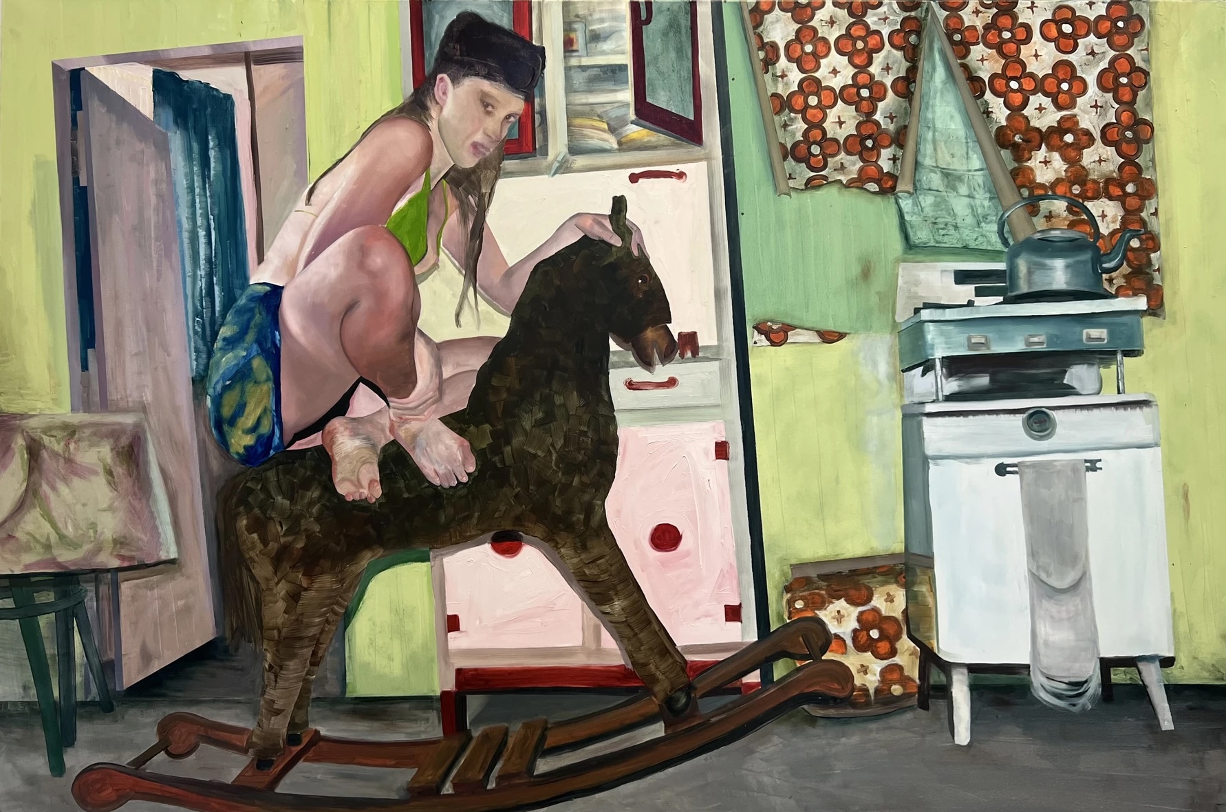 Eileen Leonard Sealy: In the ticking room | Friday 17 January  – Saturday 1 March 2025 | The LAB | Image: painting of a sideways view of a figure sitting on a dark rocking horse in a kitchen setting; the figure has long black hair and is wearing a green bikini top and a patterned skirt; legs are drawn up onto the back of the horse, and the left hand holds the top of the horse’s head while the face is turned to look at the viewer; the kitchen is fairly old-fashioned, with an old stove and cooker, an old standing press, and an isolated patch of wallpaper which is peeling dramatically above the stove; there is a door to another room behind the figure 