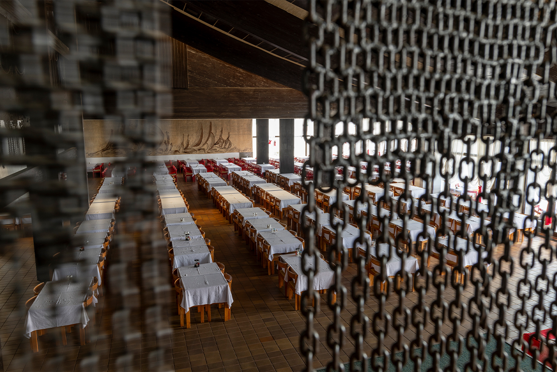 Barbara Knežević: Gvozdene Kapije (The Iron Gates), 2024, production still. Photography by Louis Haugh. | Barbara Knežević: The Iron Gates (Gvozdene Kapije) | Saturday 29 March  – Friday 30 May 2025 | Solstice Arts Centre | Image: Barbara Knežević: Gvozdene Kapije (The Iron Gates), 2024, production still. Photography by Louis Haugh | close to the camera, chains hang down from above, parted towards the middle, and partially hiding a view of a very large refectory; maybe 50 tables are lined up in rows, each with a white tablecloth and four chairs and what may be some condiments; there are supporting columns and what may be signs of a sloping ceiling; a far wall has a large hanging or mural, which appears to be depicting sailboats at sea 