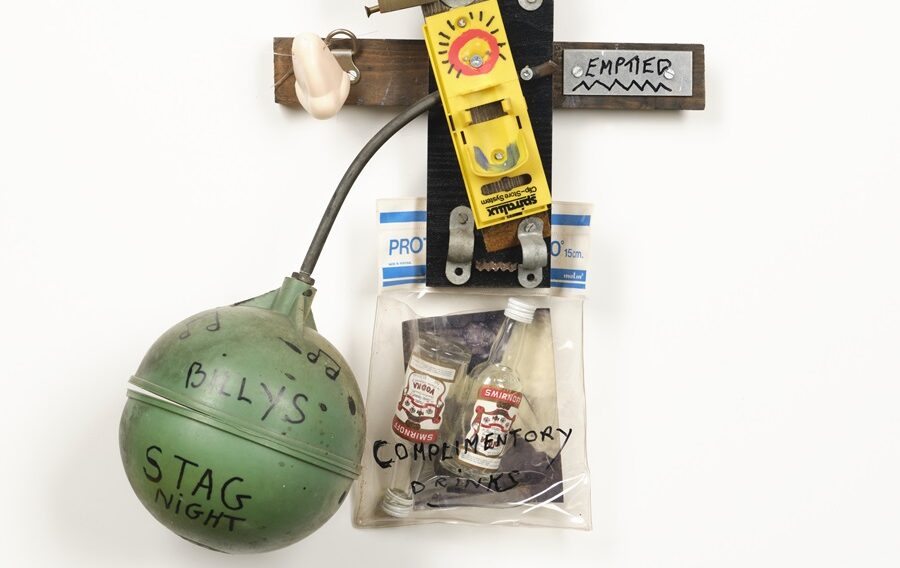 Image: Gerry Gleason, Billy the Kids Stag Night. Mixed-Media (various materials retrieved from bomb damaged building skips, Belfast city centre), 1992. Photograph by Simon Mills | Collection Highlights – Gerry Gleason | Thursday 6 February  – Saturday 8 February 2025 | Golden Thread Gallery | Image: Gerry Gleason, Billy the Kids Stag Night. Mixed-Media (various materials retrieved from bomb damaged building skips, Belfast city centre), 1992. Photograph by Simon Mills. | photo of various items nailed or screwed to a white wall, including a green ball-cock, a fake nose, two very small empty Smirnoff bottles in a plastic bag, a crossbar possibly of wood, with a label at one end that reads ‘EMPTIED’ 