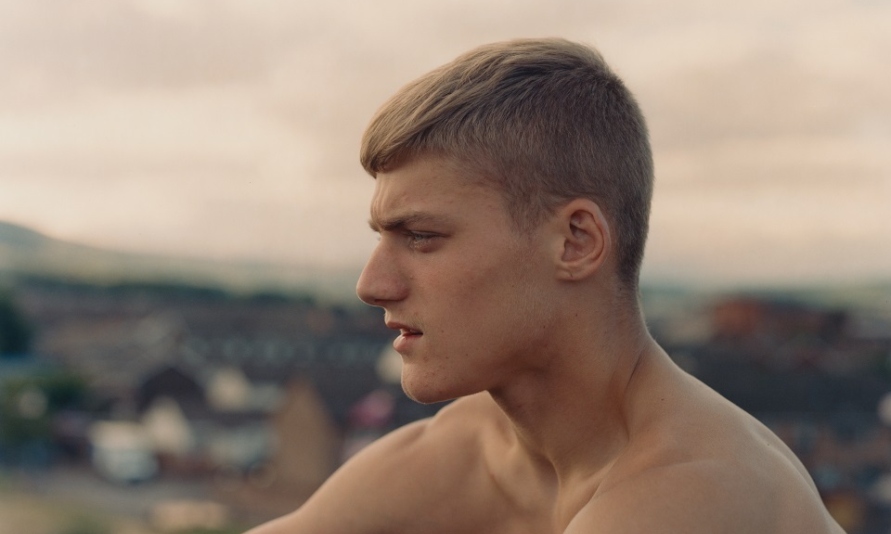 Courtesy Enda. Bowe | Enda Bowe: Love’s Fire Song | Saturday 15 March  – Saturday 19 April 2025 | The Source Arts Centre | Image: profile shot of bare-shouldered, short-cropped, frowning young adult looking towards the left edge of the image; behind is a blurred townscape plus, probably, low hilles; the sky is an orange grey 