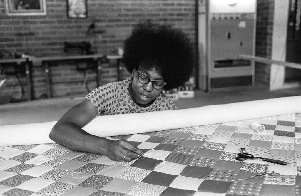 Kith & Kin: The Quilts of Gee’s Bend | Friday 28 February  – Monday 27 October 2025 | IMMA | Image: black-and-white photo of someone working on a quilt; we see just head and shoulders and right arm of the figure, with the hand probably pulling a needle through the quite, which is a patterns of squares or diamonds; behind we see what appears to be a workshop 