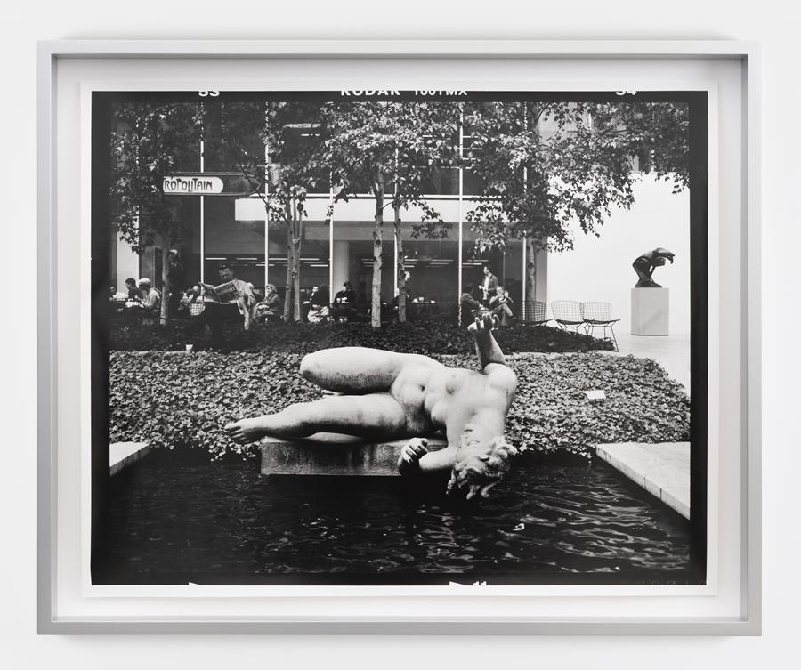 Gerard Byrne, M7 / Roll 32 / Exp 08, 2008 – ongoing, hand-printed silver gelatin print, edition of 3, 38.6 x 47.8 cm print size | Gerard Byrne: The Struggle with the Angel | Saturday 1 March  – Saturday 5 April 2025 | Kerlin Gallery | Image: Gerard Byrne, M7 / Roll 32 / Exp 08, 2008 – ongoing, hand-printed silver gelatin print, edition of 3, 38.6 x 47.8 cm print size | black-and-white photo taken in the courtyard of, possibly, of a museum of modern art, probably in a French-speaking area, judging by a sign that we can see with letters ‘opolitain’; there is a modern-style in the background, a modern-style sculpture on the right side of the image, what appears to be a café area in front of the building, with some narrow tree trunks and leaves aboe, then foliage as ground cover closer to the camera, then a pool of dark water with a pale statue in classical style, probably on its side, of a curvy naked figure with upraised harms as though fearing a fall into the water.