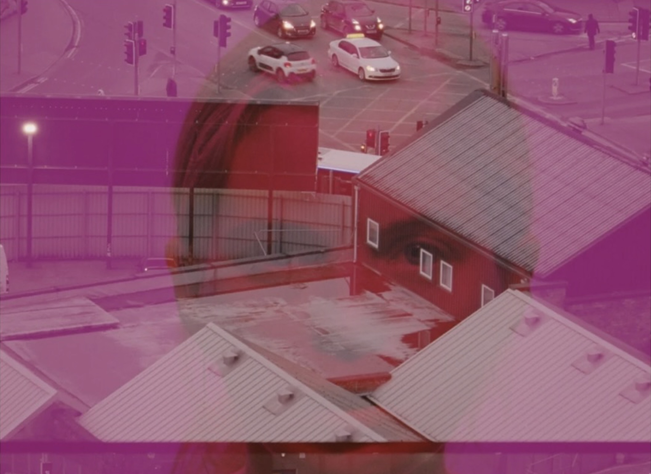 Cropped still from History of the Present; courtesy Maria Fusco and Margaret Salmon | Maria Fusco & Margaret Salmon: History of the Present | Saturday 15 February  – Saturday 29 March 2025 | Golden Thread Gallery | Image: cropped still from History of the Present; courtesy Maria Fusco and Margaret Salmon | image of head in shoulders of a figure looking at the camera; strong red tones; superimposed on this still are two others, the top one a possibly taken inside some sort of security compound – we see roofs and high walls, and beyond the walls we see a normal ciy scene of cars at an intersection; this still covers the top two-thirds of the image, with another still covering the bottom third, again above the face still; this still may have been recorded at the same location as the first – though we only see traffic 