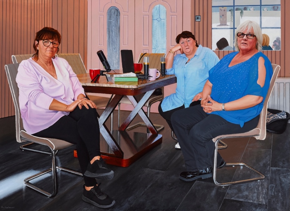 Paul MacCormaic: The Kavanagh Sisters Making Their Podcast | Paul MacCormaic: The Vanquished Writing History | Friday 21 February  – Sunday 23 March 2025 | Royal Hibernian Academy | Image: Paul MacCormaic, The Kavanagh Sisters Making Their Podcast | three figures sitting at what seems to be a marble-topped table on which are three microphones, a laptop, a few books, and three mugs of tea or coffee – two red, one pink; the figures are turned from the table, looking towards us; the chairs are modern, office-type though not swivel; the floor is slabs of polished dark stone, the decor is largely pink, with a window to the outside, which seem bright with a possible lake or se in the distance 