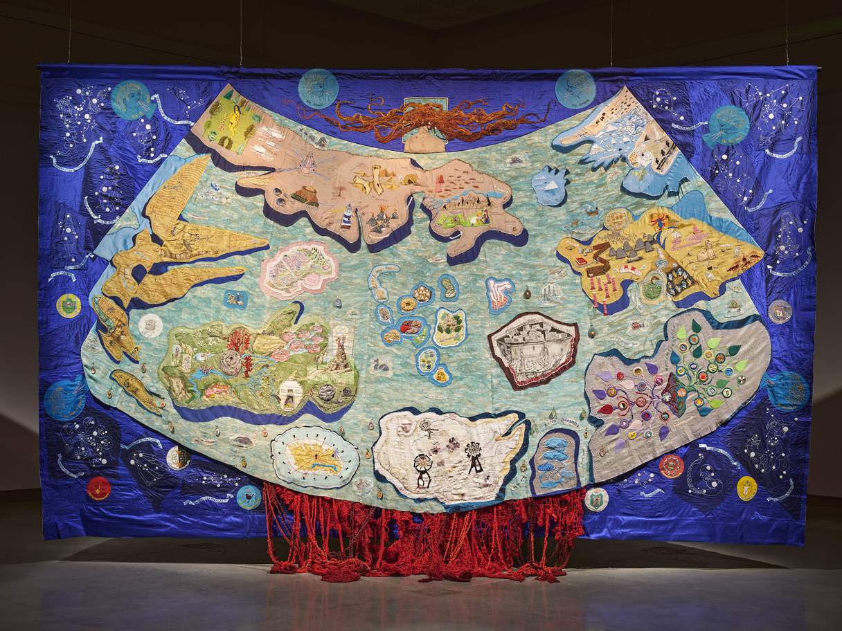 Alice Maher and Rachel Fallon, installation view of The Map at Rua Red South Dublin Arts Centre, courtesy of the artists. | Alice Maher and Rachel Fallon: The Map and The Mantle | Saturday 1 February  – Saturday 12 April 2025 | The Dock | Image: Alice Maher and Rachel Fallon, installation view of The Map at Rua Red South Dublin Arts Centre, courtesy of the artists. | a hanging tapestry dominated by a ‘map of the world’, though this world seems to have little or nothing to do with ours – unusual shapes with an illusion of 3D edges, colourful details on each landmass, the overall shape like a mantle that someone i wearing – reinforced by the appreances of the back of head and shoulders with very long wild red (orange) hair appearing above the world; the mantle would clearly be way too big for this person; around the mantle we see what may be sorts of celestial bodies; coming out at the bottom of the mantle are bulky orange-red strands of material; the whole is clearly very wide and tall, and we see it suspended in a dark space, lit from above 