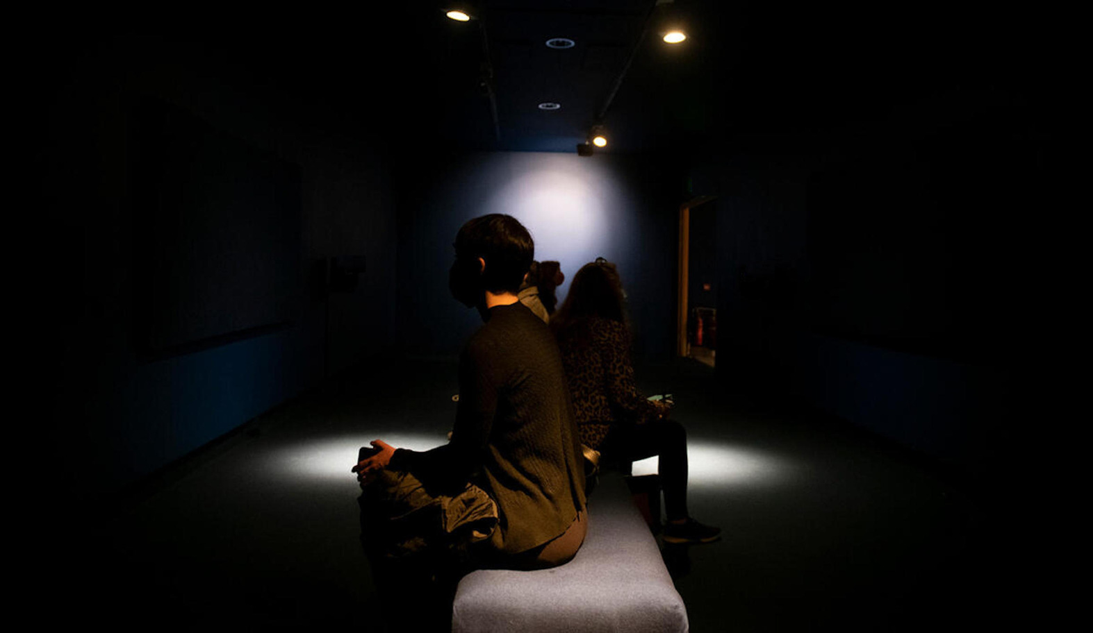 Installation view of We Are The Map by Sinéad Gleeson and Stephen Shannon at Rua Red South County Dublin Arts Centre. | Sinéad Gleeson & Stephen Shannon: We Are The Map | Saturday 1 February  – Saturday 12 April 2025 | The Dock | Image: Installation view of We Are The Map by Sinéad Gleeson and Stephen Shannon at Rua Red South County Dublin Arts Centre | photo: side-one view of a few people sitting in d dark space with very sparse lighting; the figure closest to us is looking to the left, at what we don’t know; cropped dark hair, dark top, left hand visible on left knee, pool of light on the floor beyond the left hand; hint of maybe two more visitors in this space; also hint of some sort of rectangular object hanging in a dark corner; four light sources visible in the ceiling 