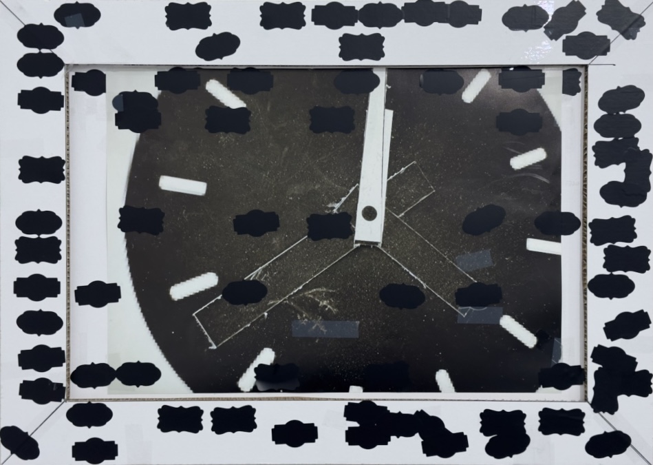 Glenn Fitzgerald, Nothing From Something, 2025, Inkjet print, tape, label stickers on honeycomb card panel, 70 x 50 cm, Image courtesy of the artist | Glenn Fitzgerald: Lucky Charms | Thursday 27 March  – Sunday 20 April 2025 | Royal Hibernian Academy | Image: Glenn Fitzgerald, Nothing From Something, 2025, Inkjet print, tape, label stickers on honeycomb card panel, 70 x 50 cm, Image courtesy of the artist | depicts a framed image of a black clock-face with white hour markings and white hands; we do not see the full circle of the face, as it is cropped by a white frame and an inner passpartout; the hands point to just after noon / midnight; there are signs of two other hands, but they are just long black rectangles with white outlines against the black of the clock face; the frame is white and simple, but it and the ‘glass’ (or whatever it is) of the clock image are part-covered by black labels that contain no writing or other information; they are fancily shaped 