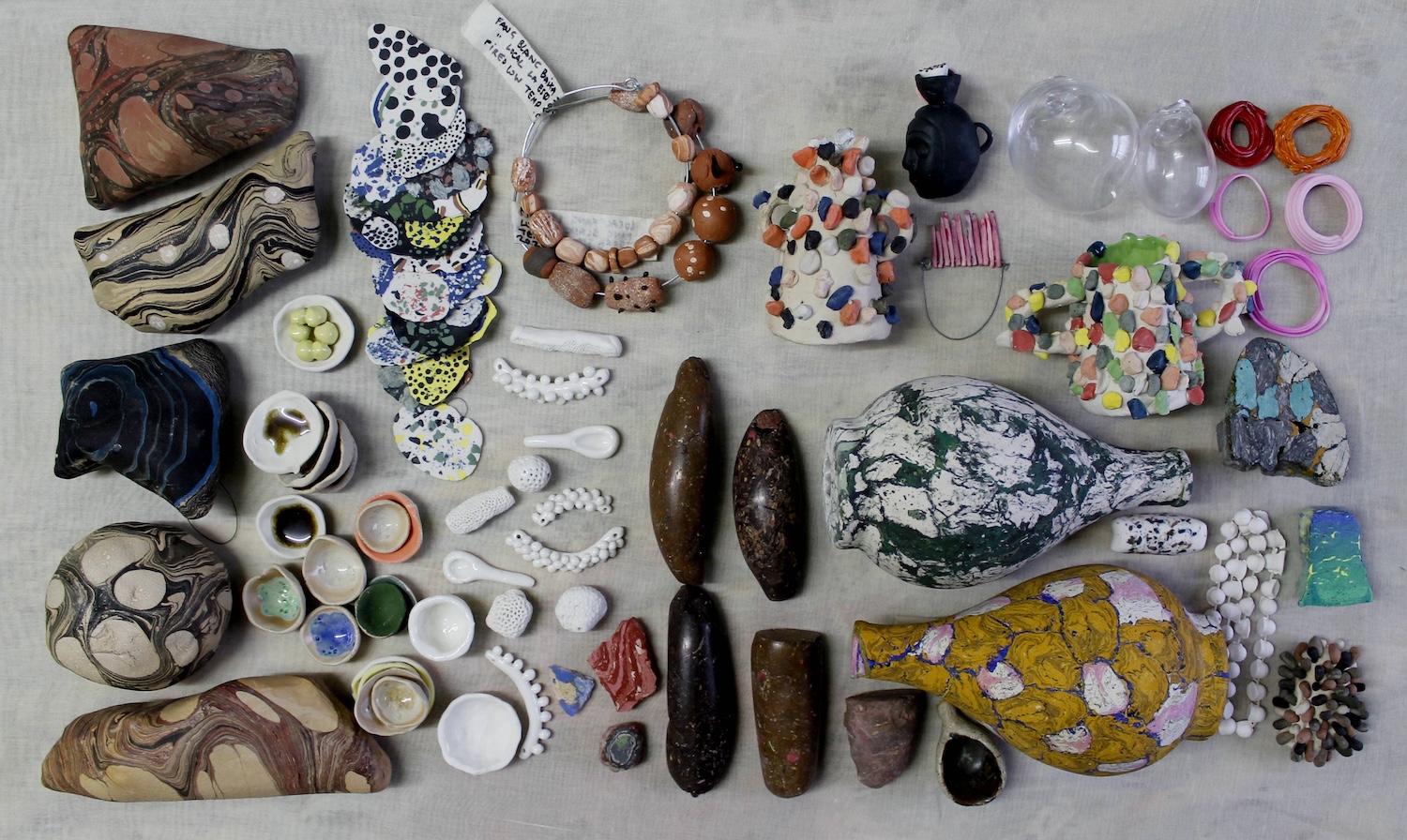 Vanessa Donoso López: A rock can hurt you, a rock can open a nut | Sunday 13 April  – Sunday 22 June 2025 | Municipal Gallery | Image: photo from above of various objects, some of which my be rocks / stones; there is a pair of bracelets top middle, for example, that seems to employ stones, but the label attached –  in Catalan and English – suggest that they were “fired low temp”; the are small white spoons, polished stones, what seems to be transparent plastic shapes that are somewhat biomorphic, loops of stuff, etc. 