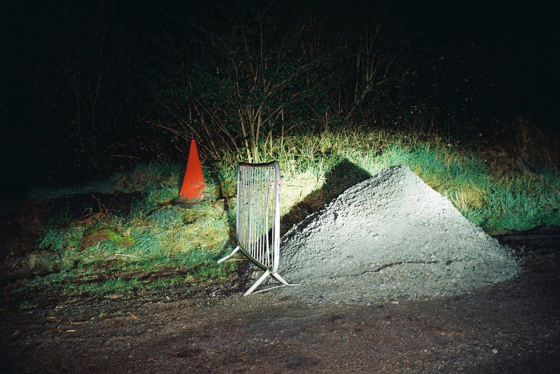 Feá McCoy: Untitled, 35mm colour negative film photograph, 2024 | Feá McCoy: What Remains Absent | Saturday 29 March  – Friday 30 May 2025 | Solstice Arts Centre | Image: Feá McCoy: Untitled, 35mm colour negative film photograph, 2024 | a photo taken at night; a bright light is shining on a mound of gravel (?) by the side of a road (perhaps); a metal crowd barrier stands to the left of the mound, and behind both we see a bright red traffic cone among grass, and other plantlife, and behind ti all, darkness