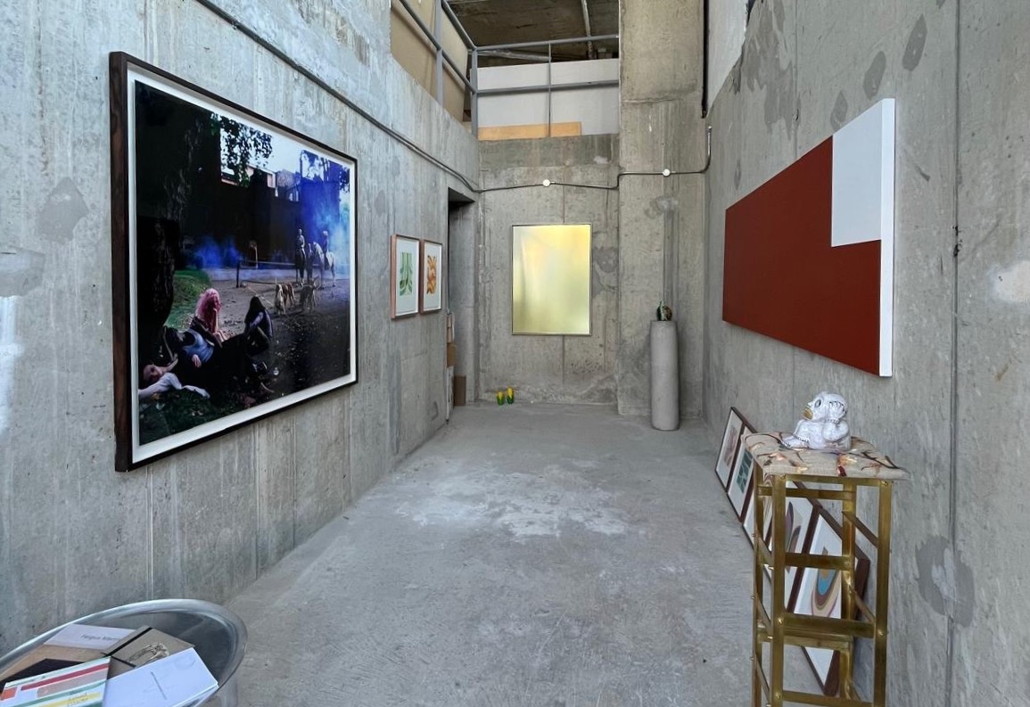 Ground | Monday 14 April  – Friday 16 May 2025 | Green On Red Gallery | Image: In what is possibly a (brutalist) side passage at Green on Red, this photo shows various artworks – a large photographic work on the left featuring people, horses with riders, and smoke, a mostly gold framed image at the end of the passageway, a concrete-looking round plinth with possibly a ceramic on it, a wide-but-not-tall, mostly redding-brown canvas facing the large photo, some framed images on the floor, some golden shelving, etc. 