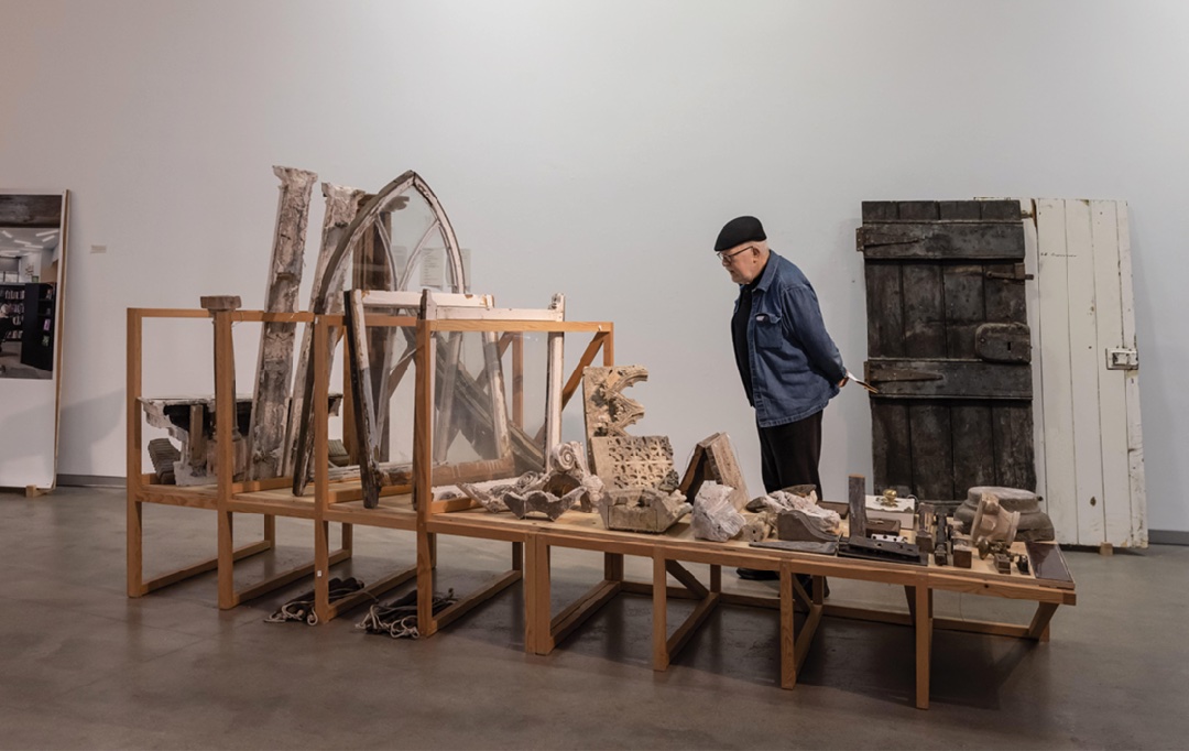 Valerie Mulvin: The Reason of Towns, installation shot, The Source, Thurles. Photo by Ste Murray. | Valerie Mulvin: The Reason of Towns | Saturday 12 April  – Wednesday 21 May 2025 | Uillinn: West Cork Arts Centre | Image: Valerie Mulvin: The Reason of Towns, installation shot, The Source, Thurles. Photo by Ste Murray. | cap-wearing gallery visitor is looking at a large selection of what are presumably everyday artefacts that can be / could be found around a town; these include a pair of white-framed sash windows and a fancier Gothic-art-type window; the artifacts are held in an open wooden frame; behind the viewer and leaning against a wall are two old doors 