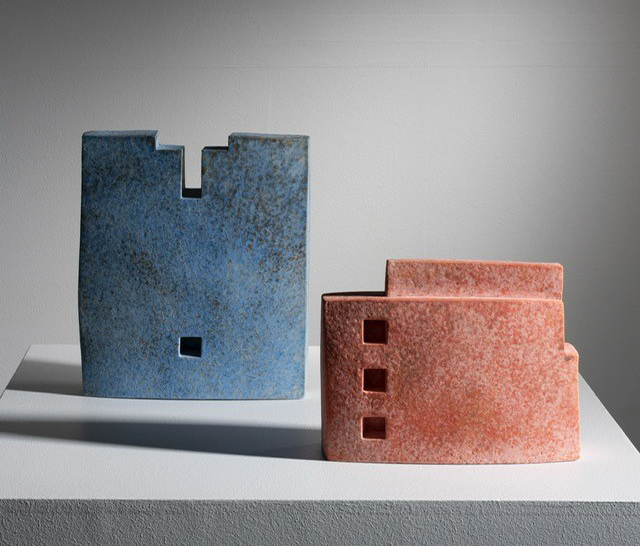 Mike Byrne: URBANESQUE | Friday 11 April  – Sunday 11 May 2025 | Limerick City Gallery | Image: photo of two ceramic works; each resembles a building, the left, blue-ish one perhaps a Norman castle with parapet and a single window low in its façade; the reddish work on the right is more like an apartment block, lower, with three square window shapes on the left, one above the other 