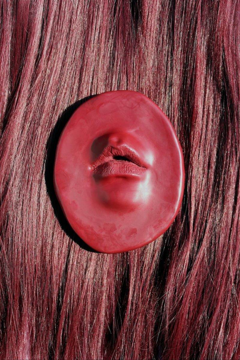 Eibh Gordon: Reliquary, as Oubliette | Thursday 1 May  – Thursday 26 June 2025 | Atypical Gallery | Image: photo; we see what seems to be a realistic mouth in a red substance that resembles plasticine; we see a bit of the upper lip as well, and a suggestion of chin; the backround apparently someone’s reddish hair 